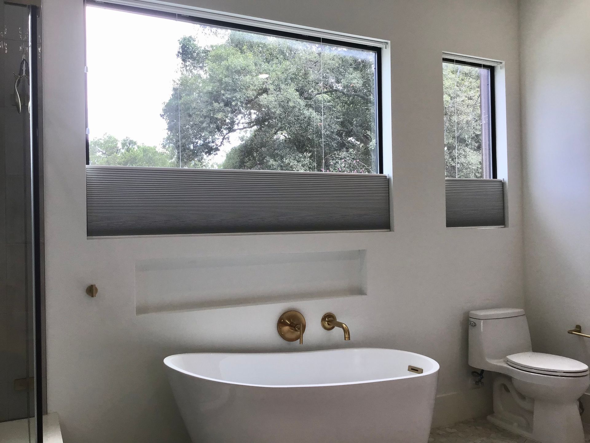 A modern bathroom featuring a white freestanding tub, gold faucet, wall niche, and two windows with grey roman shades.