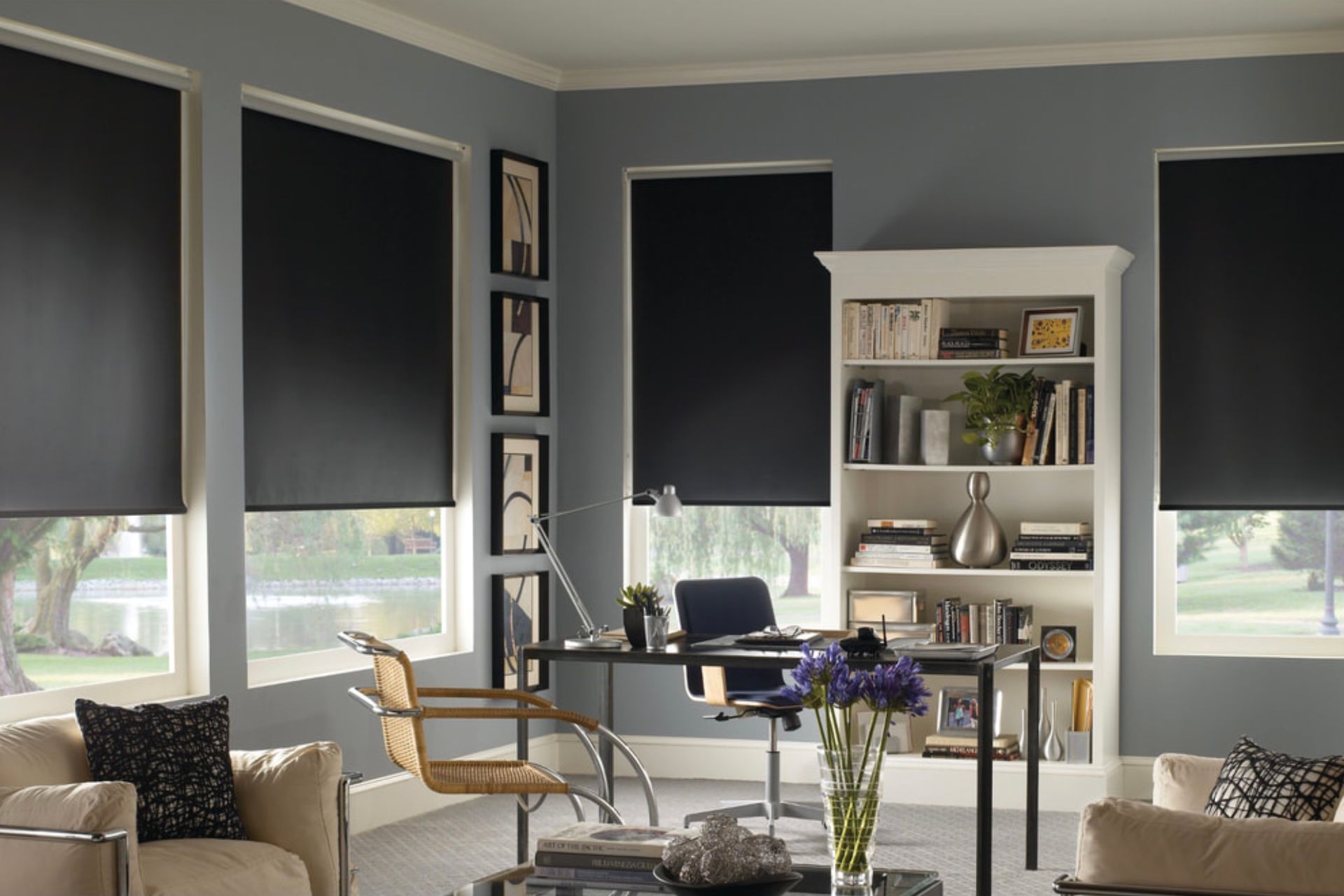 A modern home office with grey walls, white bookshelves, and dark roller shades on three windows.