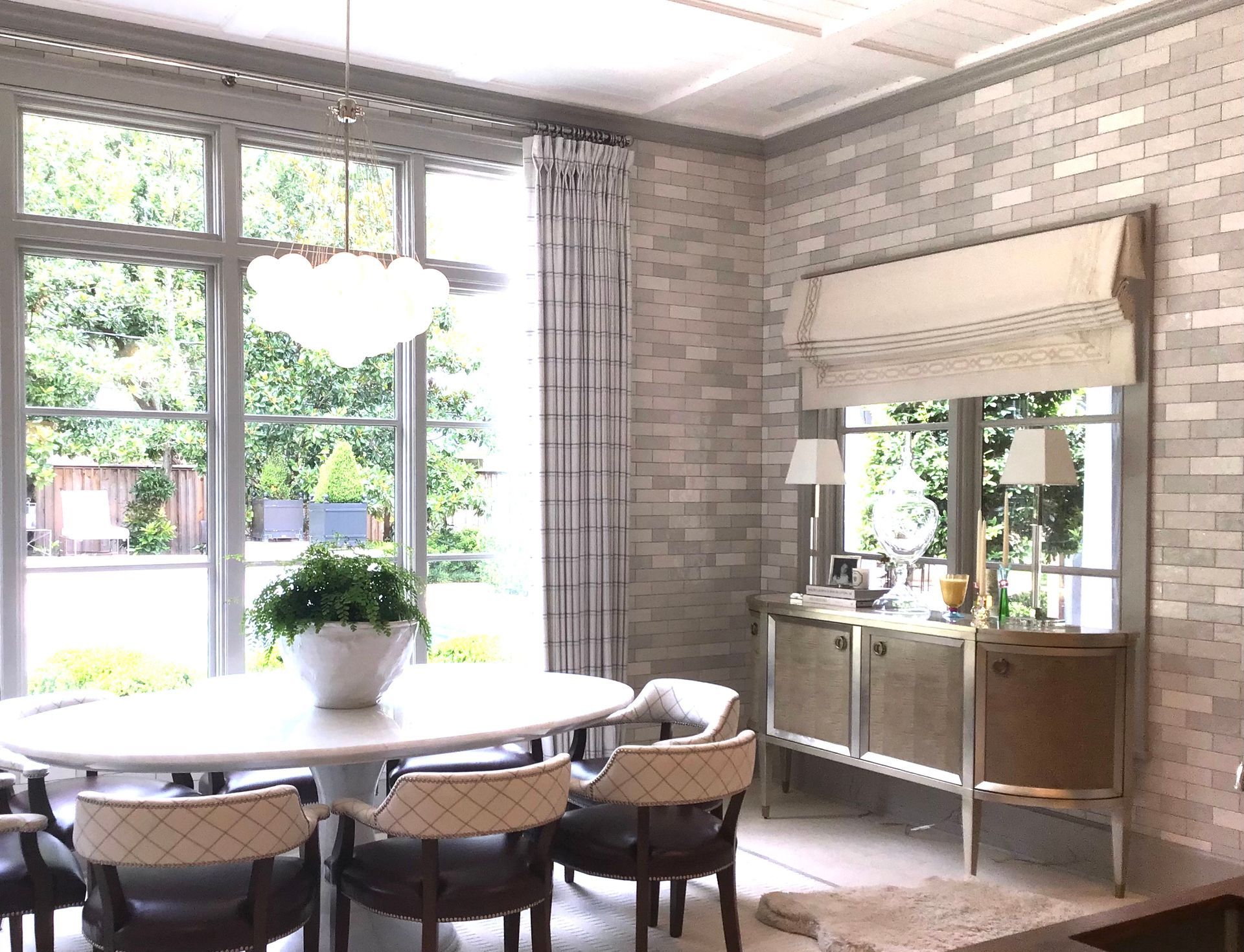 A dining room with a white oval table, upholstered chairs, a chandelier, and a sideboard against a light brick wall.