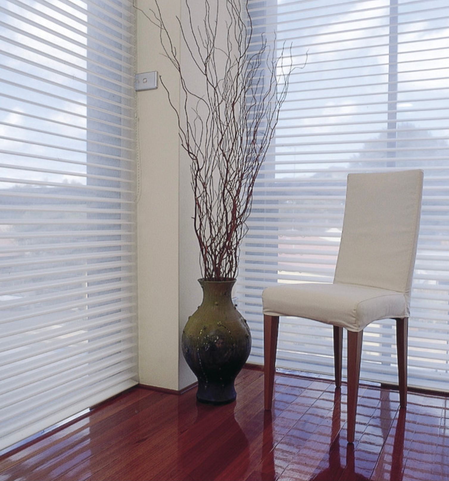 A white dining chair sits on a wood floor next to a tall, decorative vase filled with thin branches against window blinds.