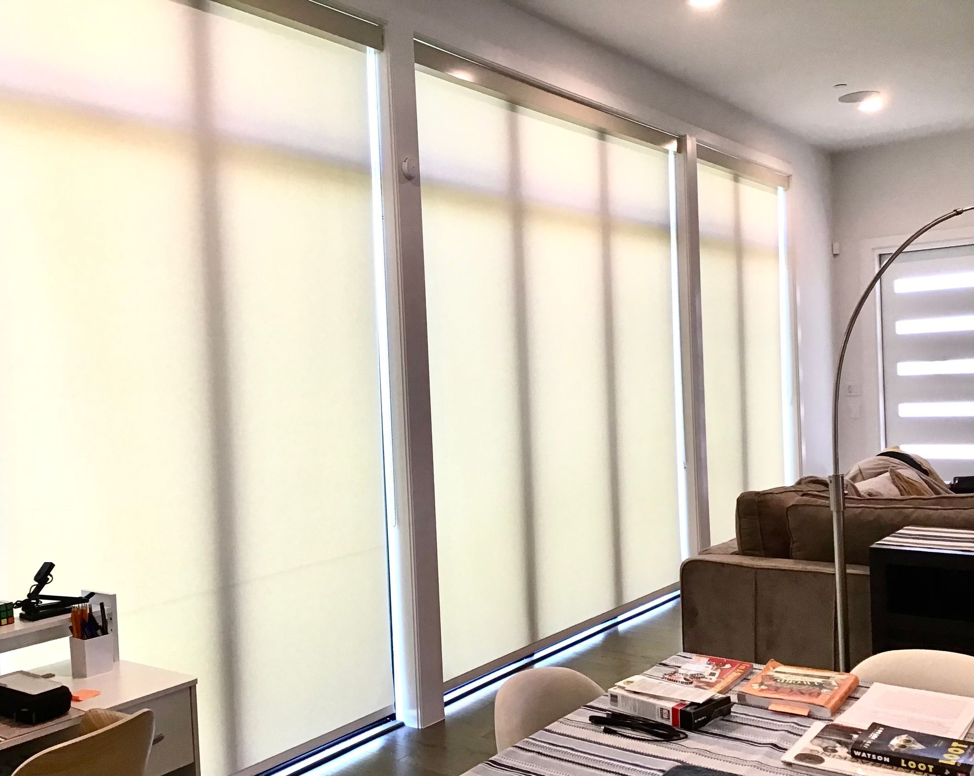 Floor-to-ceiling windows with cream-colored roller shades in a modern living space with a dining table and sofa nearby.