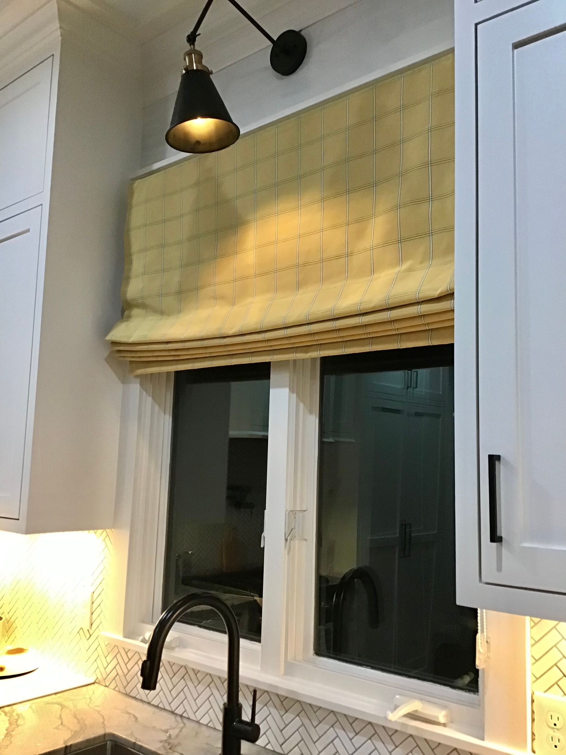 A kitchen sink window with a yellow patterned roman shade and a matte black wall sconce above.