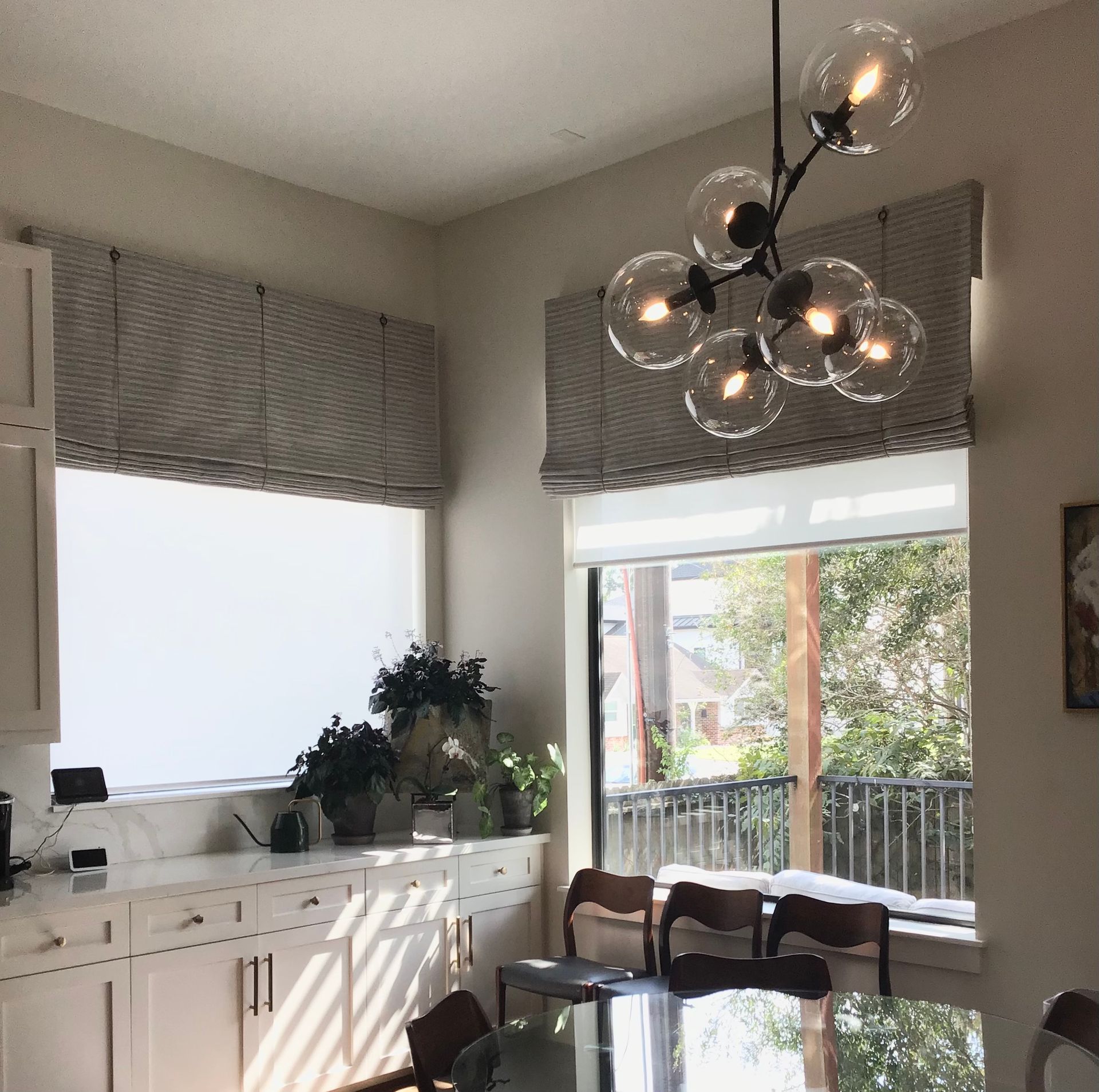 A bright dining area features a modern glass globe chandelier hanging above a table, framed by windows with textured shades.