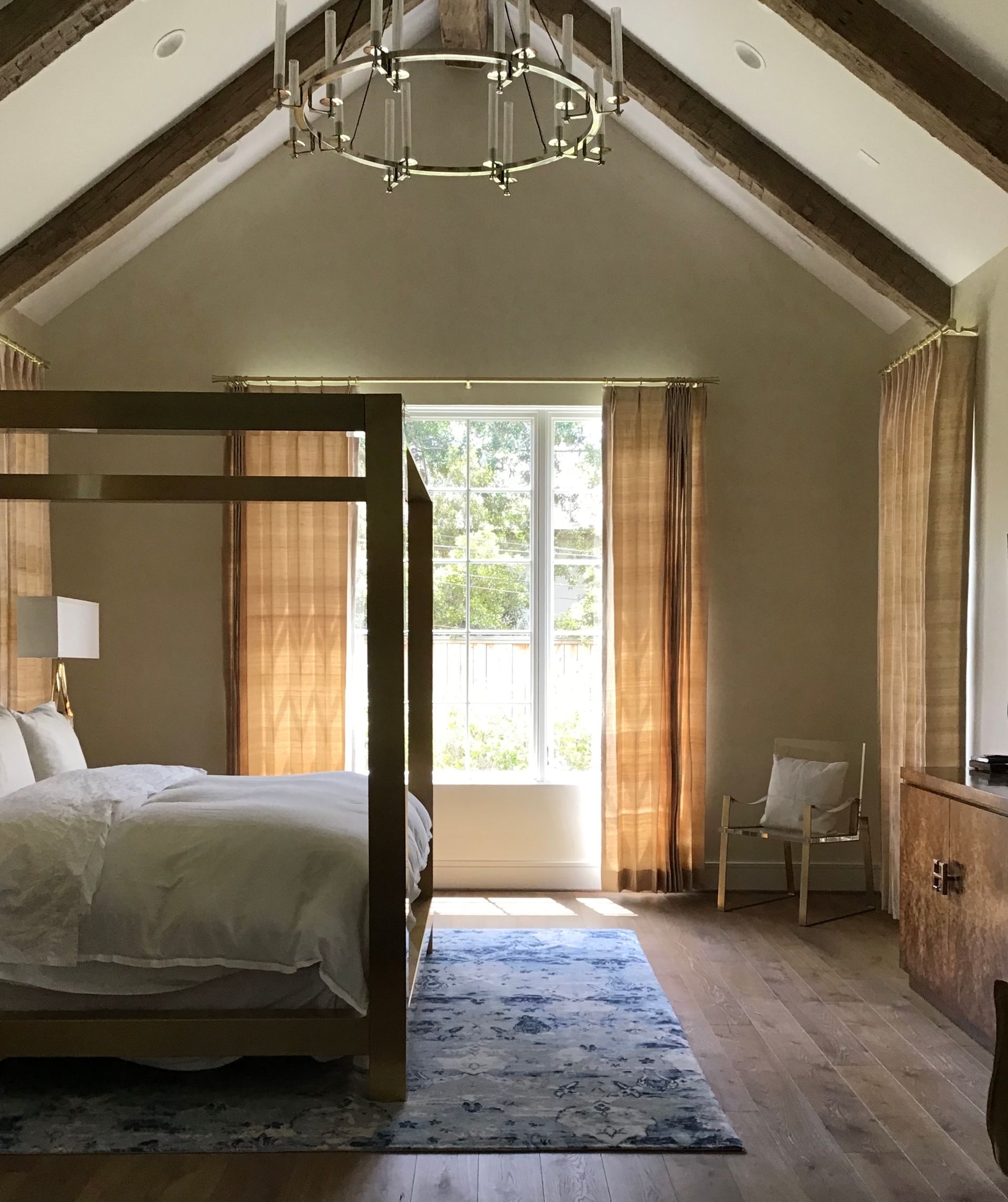 A bedroom with a wooden canopy bed, vaulted ceiling, rustic wood beams, a chandelier, and light-filled windows with drapes.