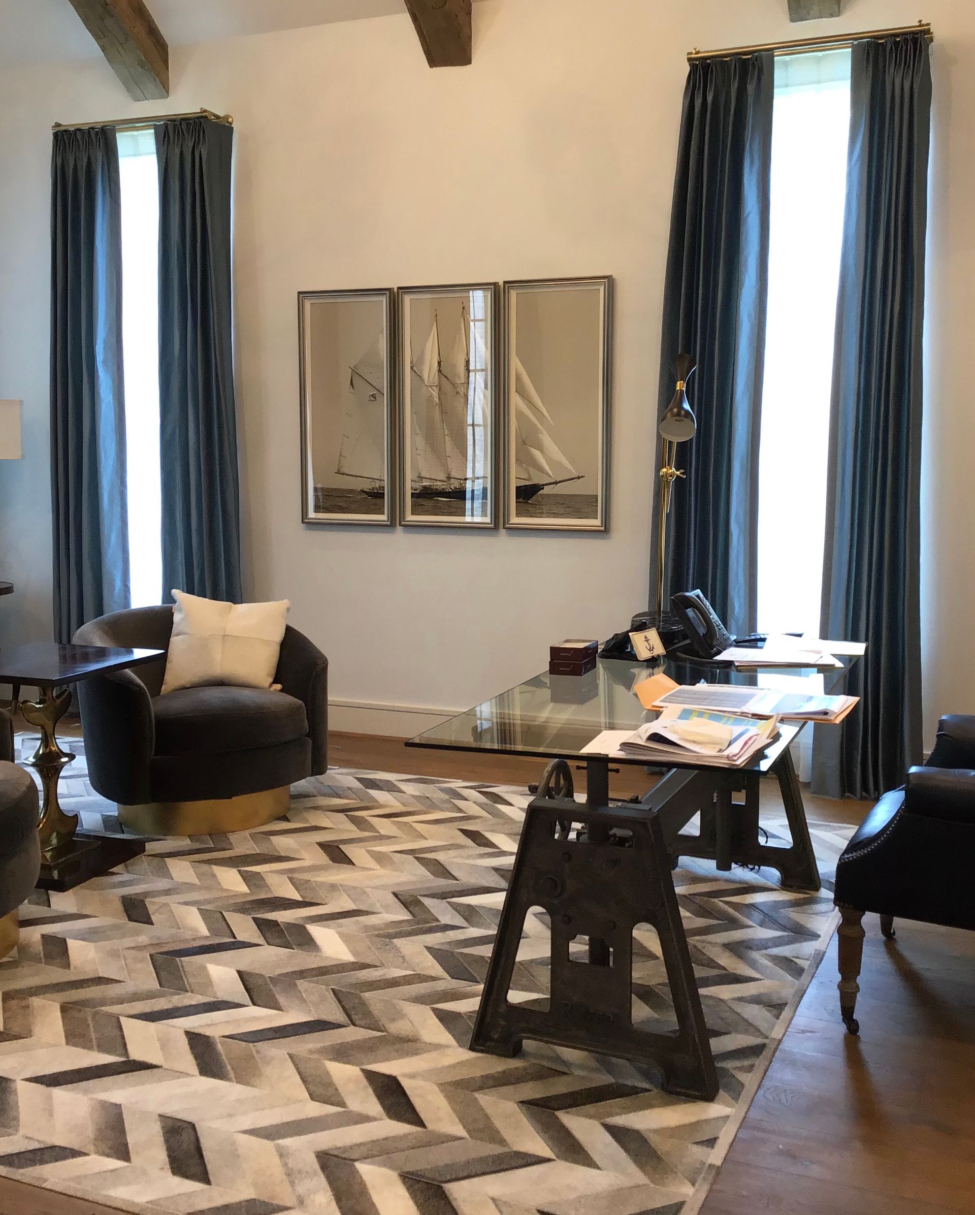 A modern home office with a glass desk, grey chevron rug, dark velvet chairs, and three sailboat prints on the wall.