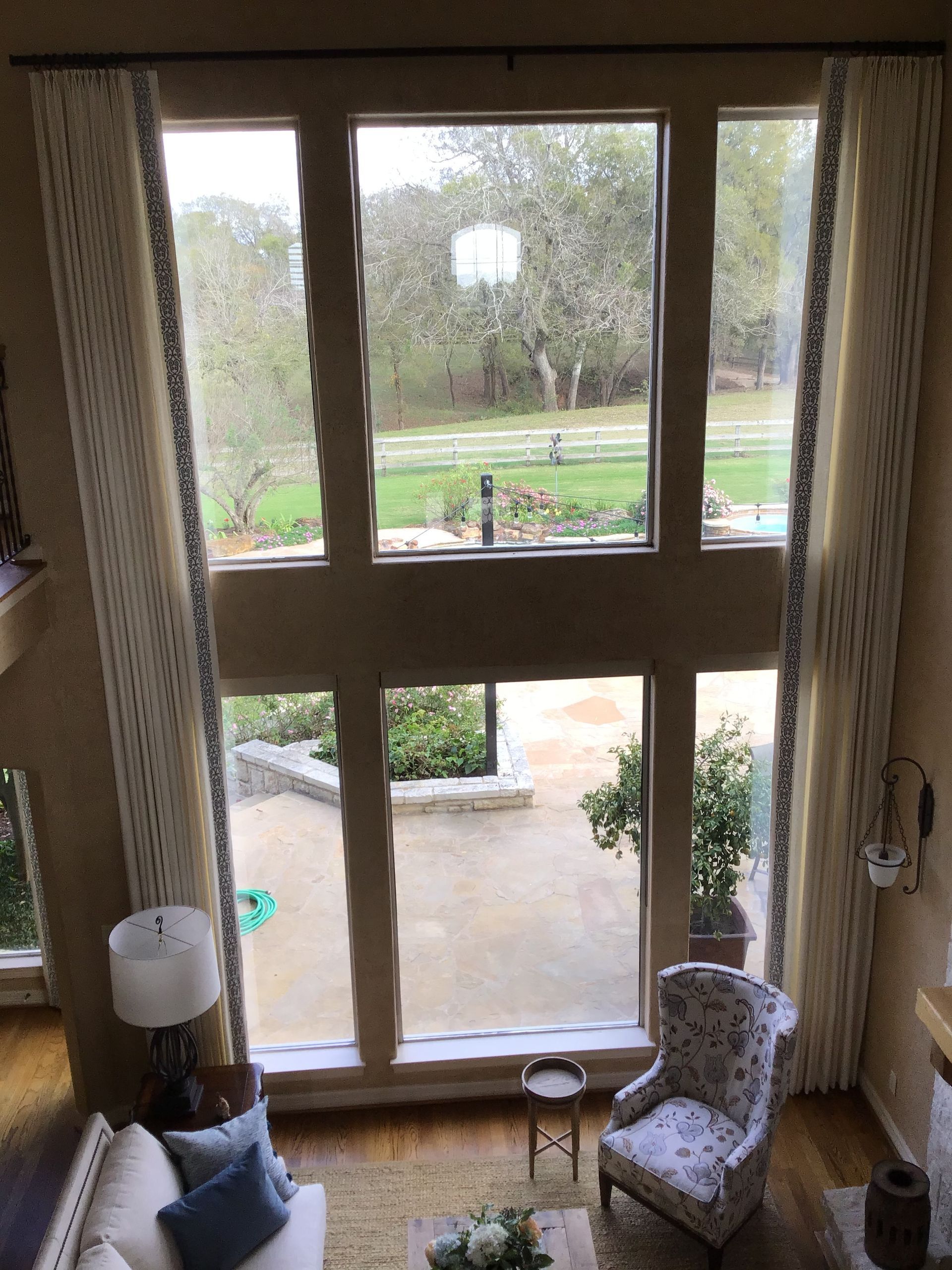 A high-ceilinged room with a large, two-story window looking out onto a backyard, framed by long, cream-colored curtains.
