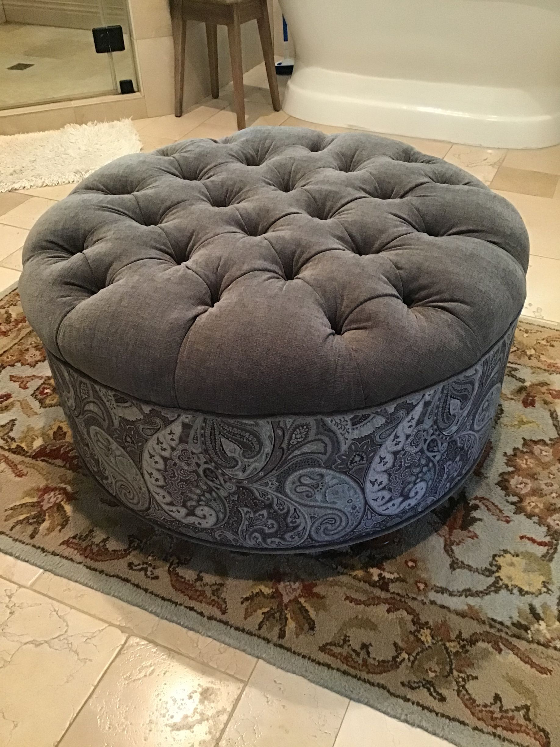 A round, tufted grey ottoman with a patterned fabric base, sitting on a decorative area rug in a room.
