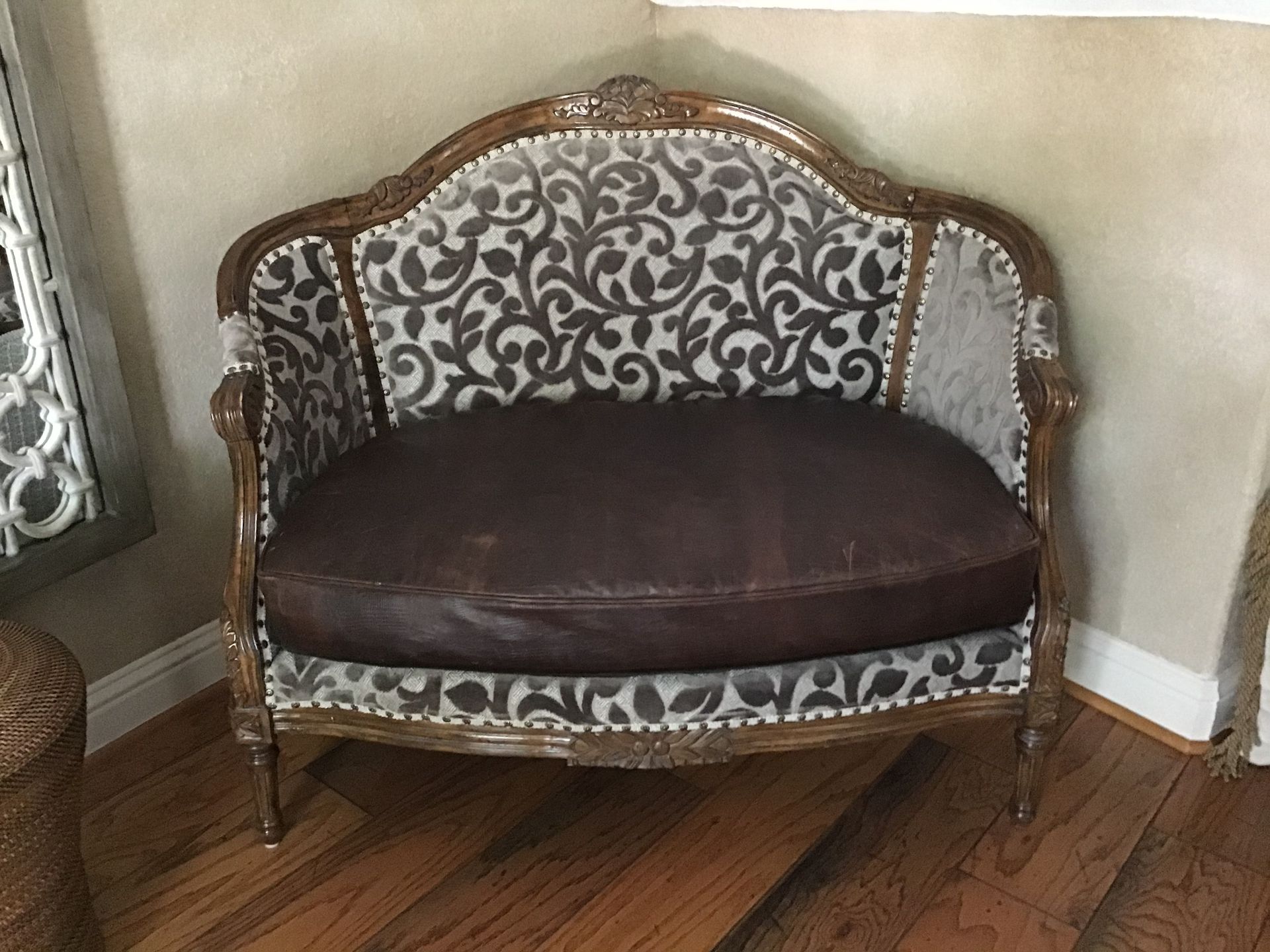 An ornate wooden loveseat with a dark brown leather seat and a gray and white leafy patterned upholstered back.