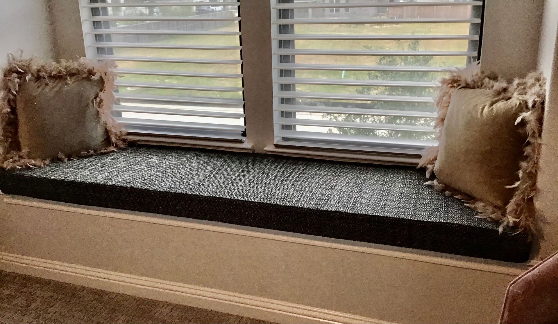 Window seat with a dark, textured cushion and two fringed beige pillows in front of closed white blinds.