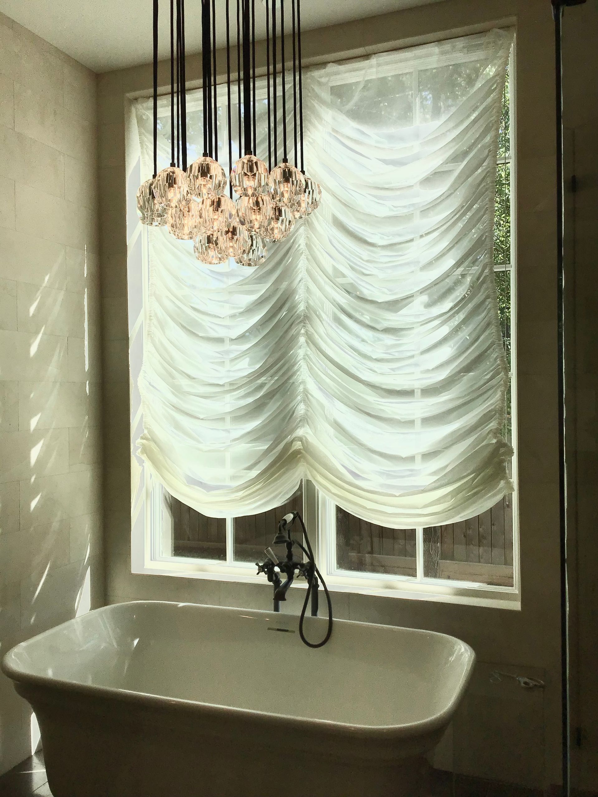 A white bathtub stands under a large window with draped, white curtains and a hanging multi-globe light fixture.