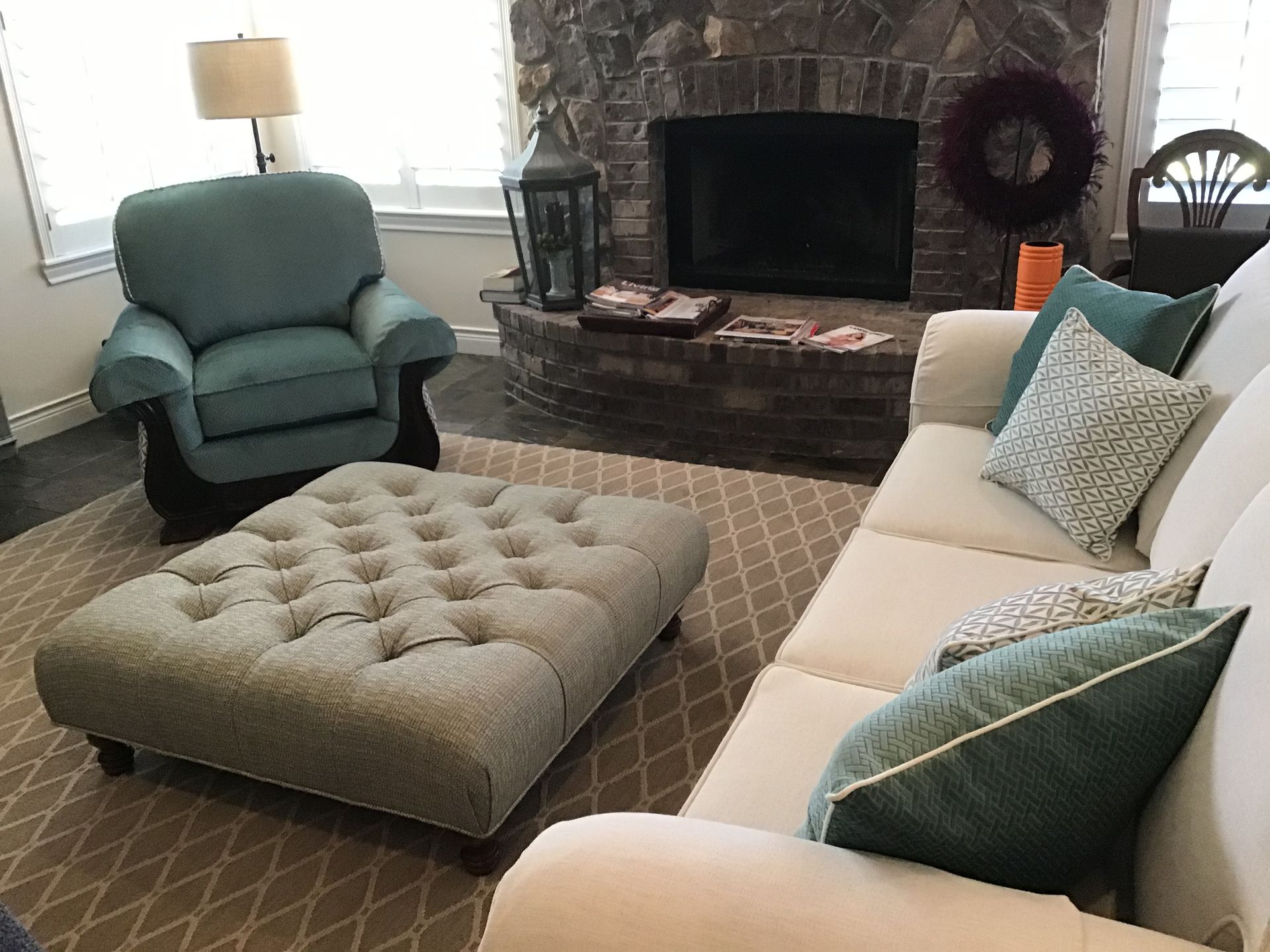 A teal armchair and white sofa with matching pillows face a stone fireplace with a tufted ottoman in the center.