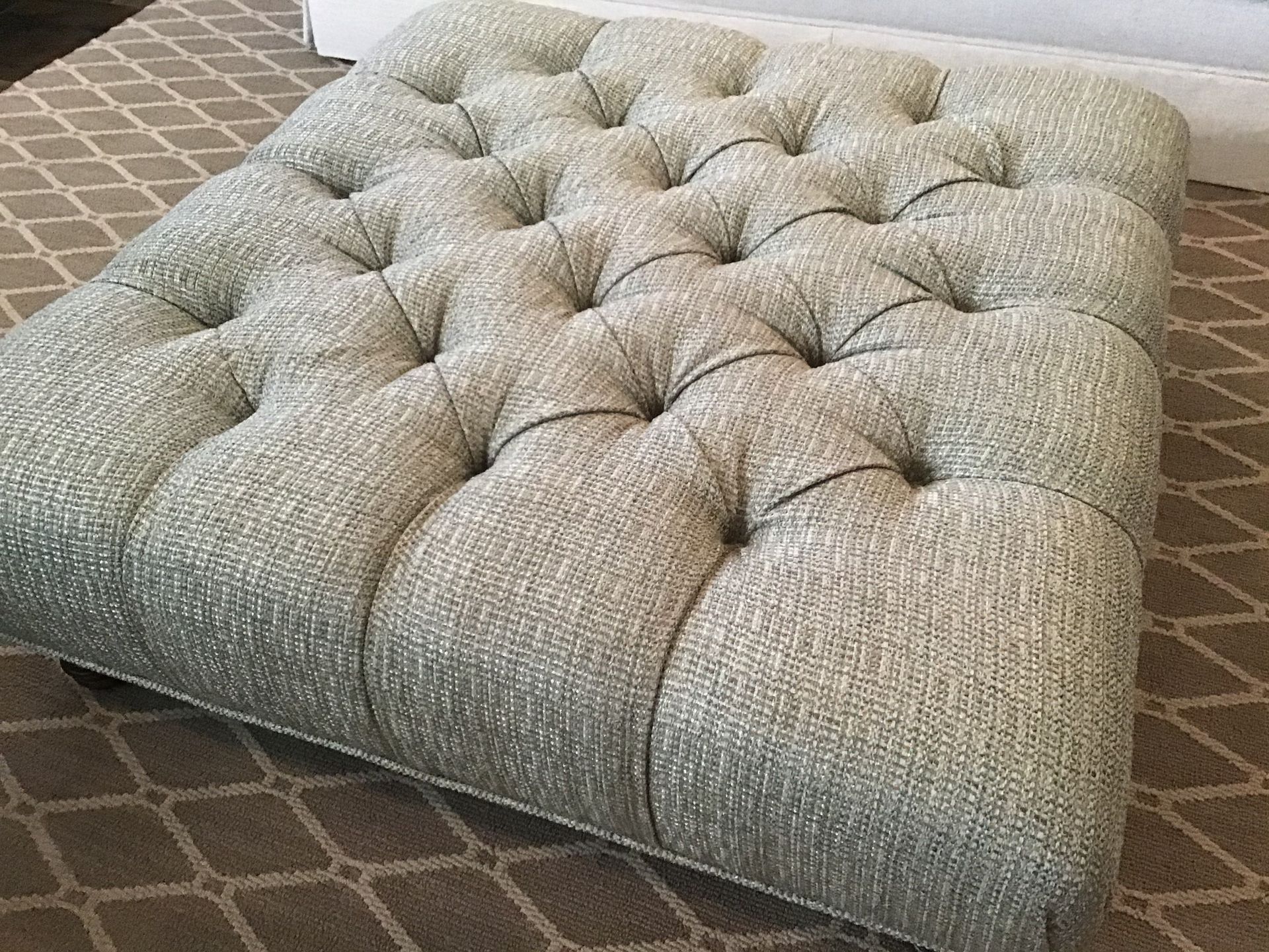 A square, tufted ottoman upholstered in light green textured fabric, sitting on a patterned brown rug.