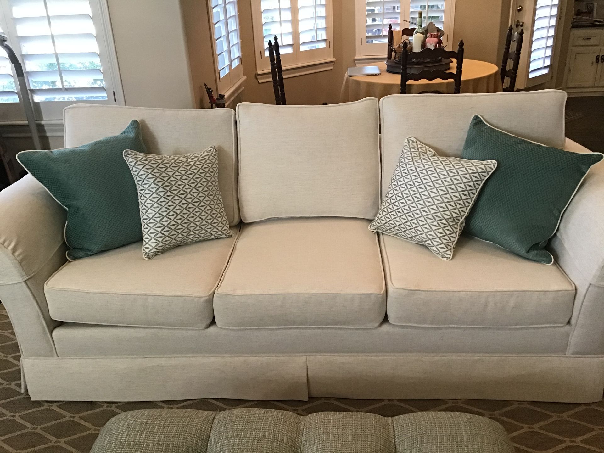 A light-colored three-cushion sofa featuring two dark teal pillows and two patterned throw pillows in a bright room.