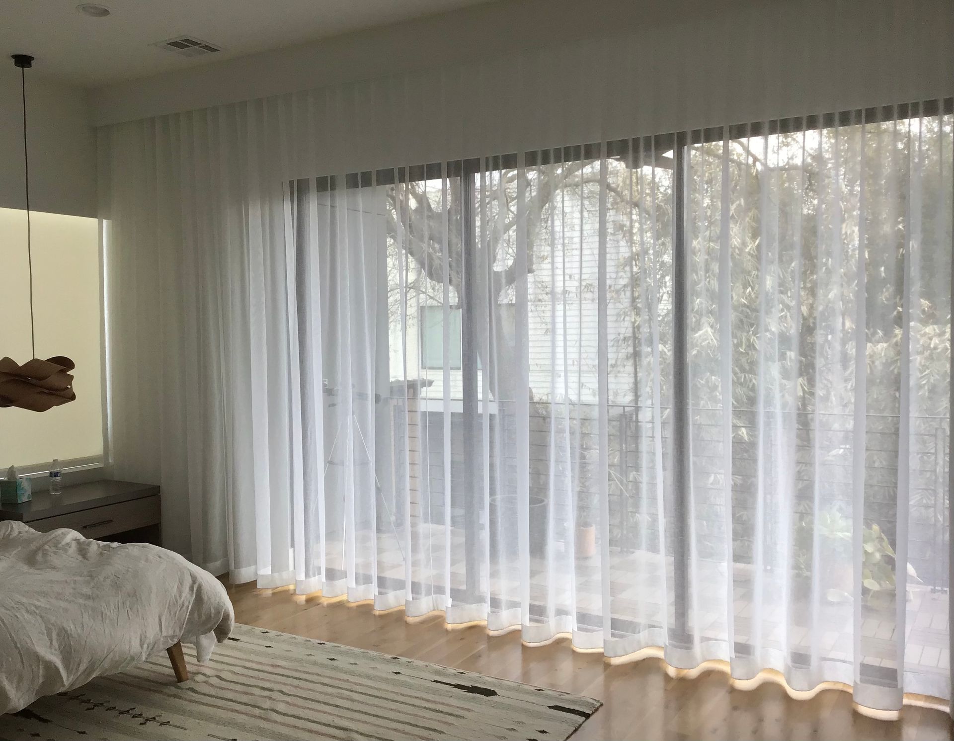 A bedroom featuring floor-to-ceiling sheer white curtains covering a large window with a view of trees.
