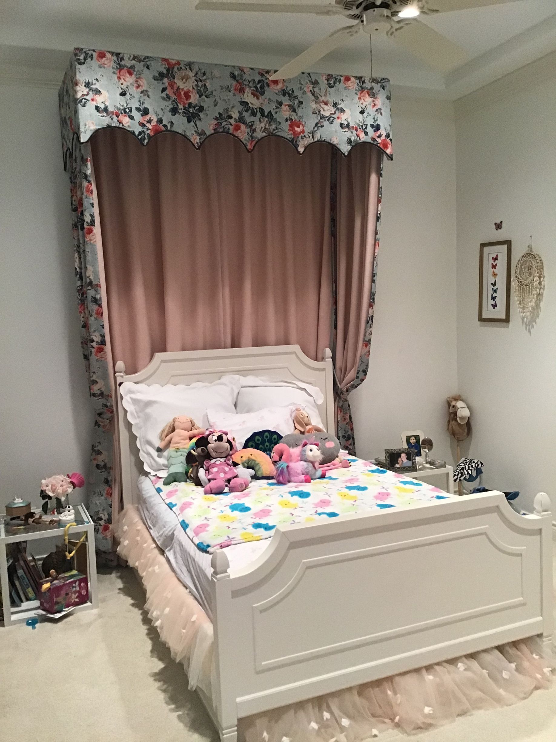 A white bed with a patterned canopy and pink curtains, featuring a colorful toy-covered quilt, in a bedroom setting.