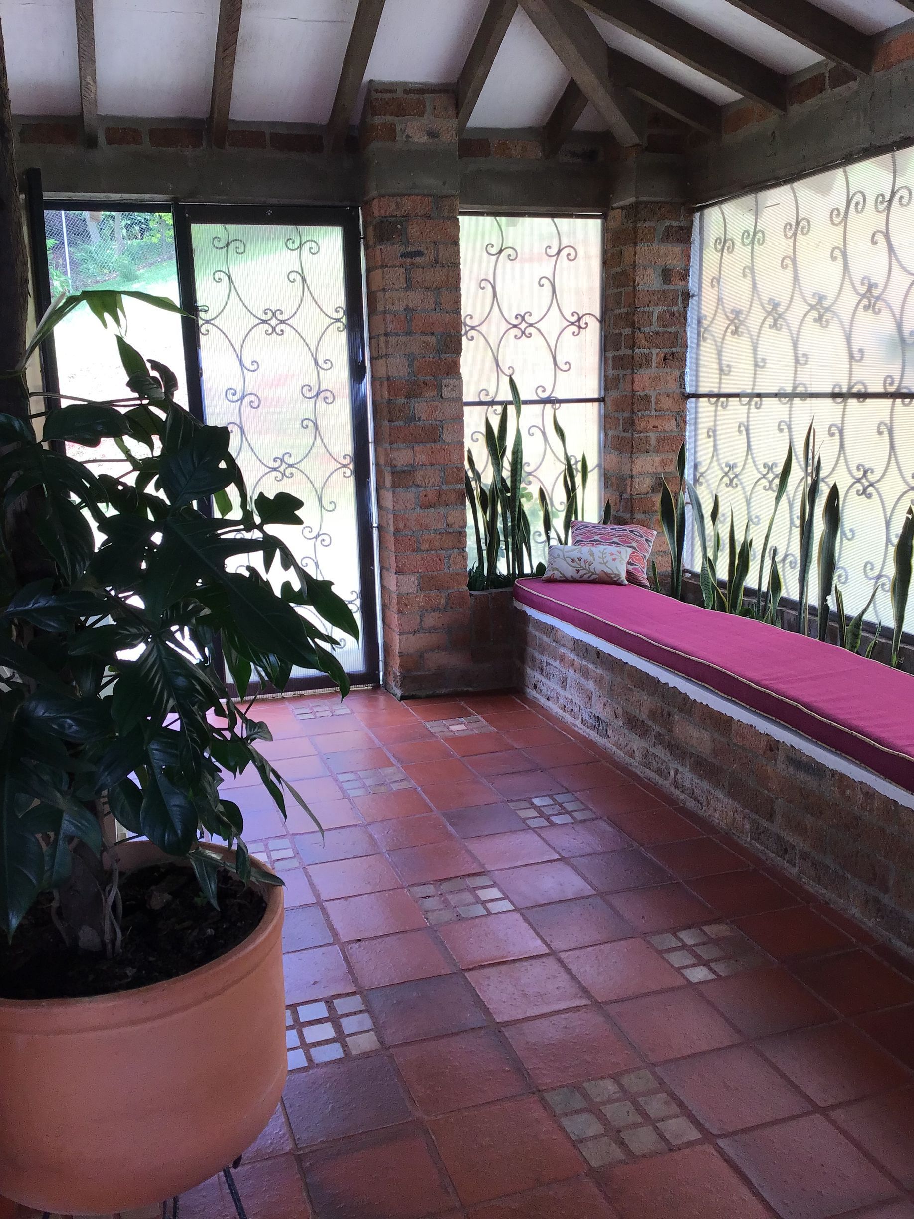 A sunroom with terracotta tile flooring, exposed brick pillars, a long bench with a red cushion, and a potted plant.