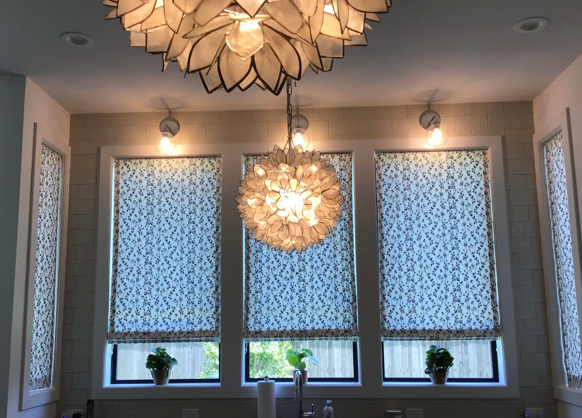 Three windows with patterned Roman shades feature two decorative hanging light fixtures and small potted plants.