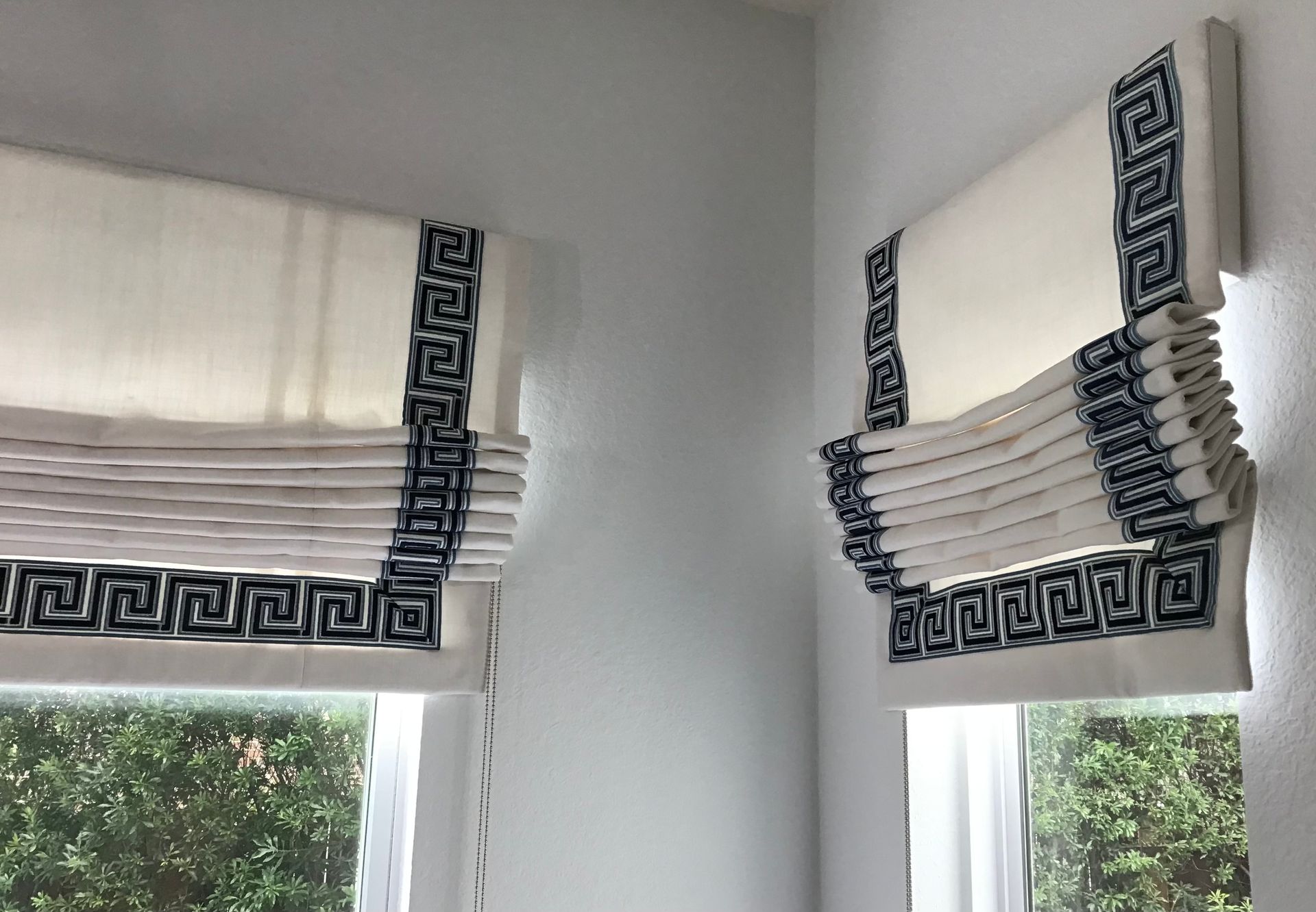 Two white roman shades featuring a dark blue Greek key pattern trim, mounted over windows on a light-colored wall.