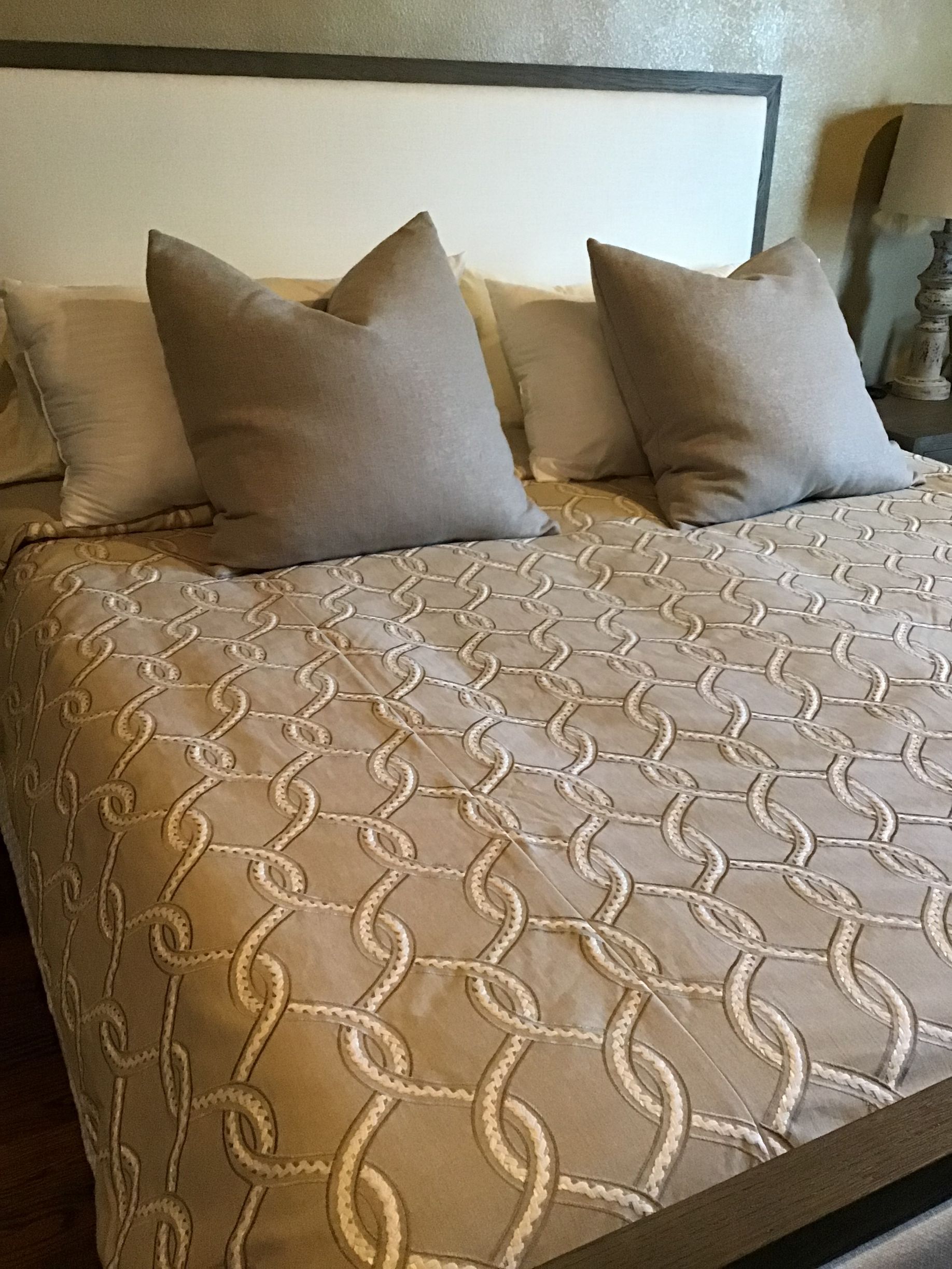 A bed featuring a tan comforter with a white chain-link pattern, topped with two large tan pillows and two smaller ones.