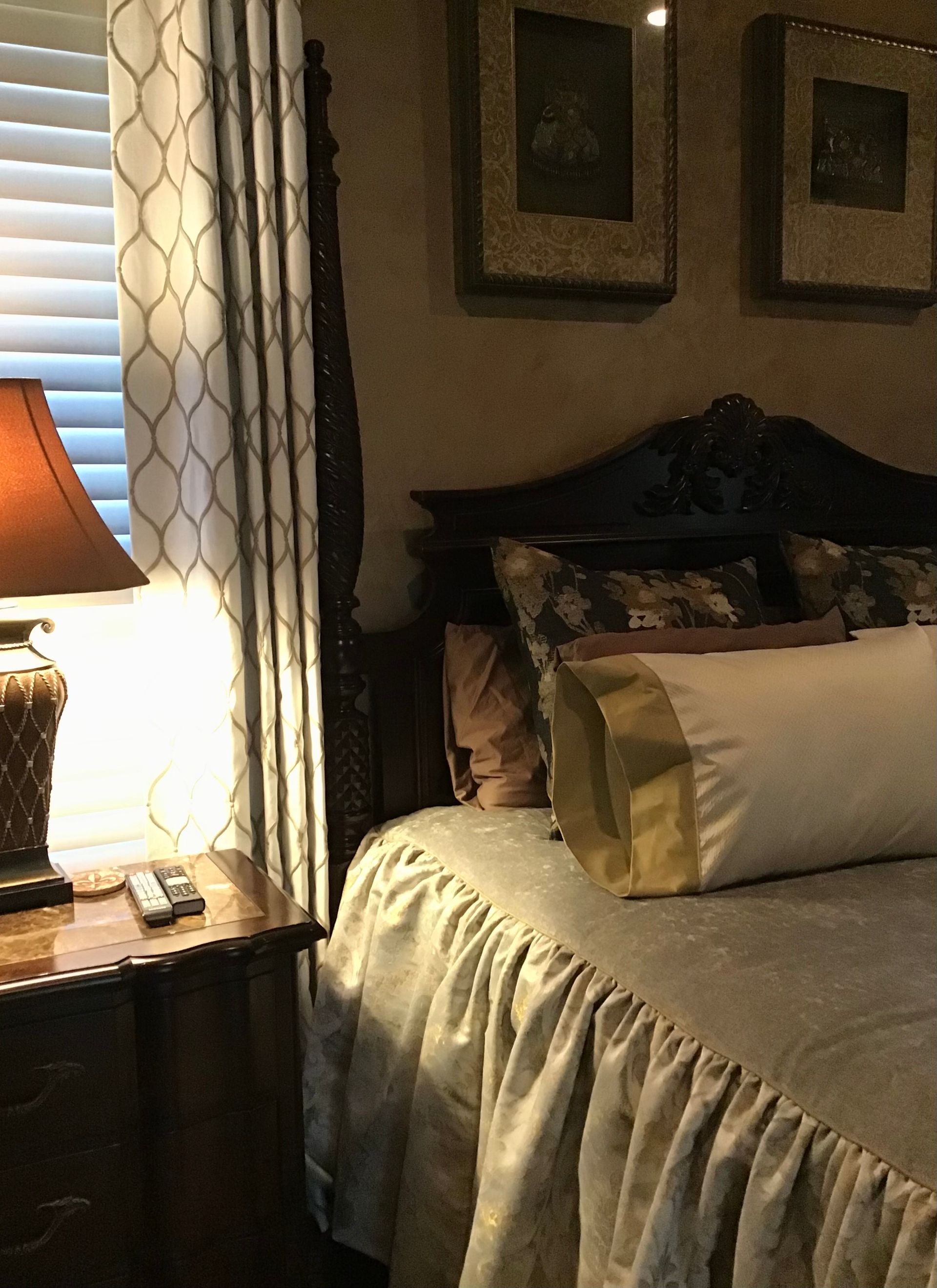 A bedroom corner with a wooden nightstand, a lit table lamp, patterned curtains, a bed, and two framed wall pictures.