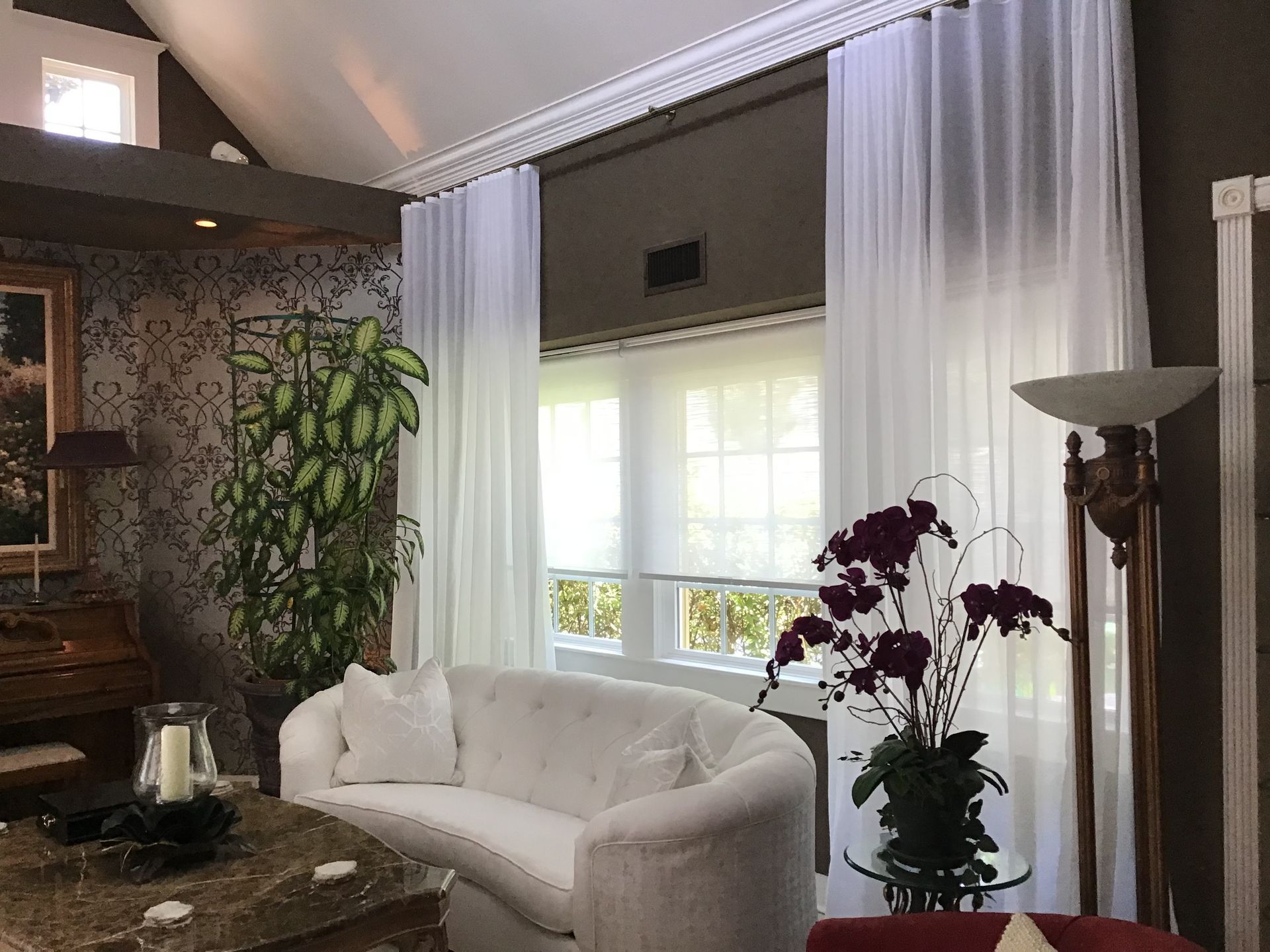 A bright living room features a white curved sofa, sheer floor-to-ceiling curtains, a large potted plant, and an orchid.