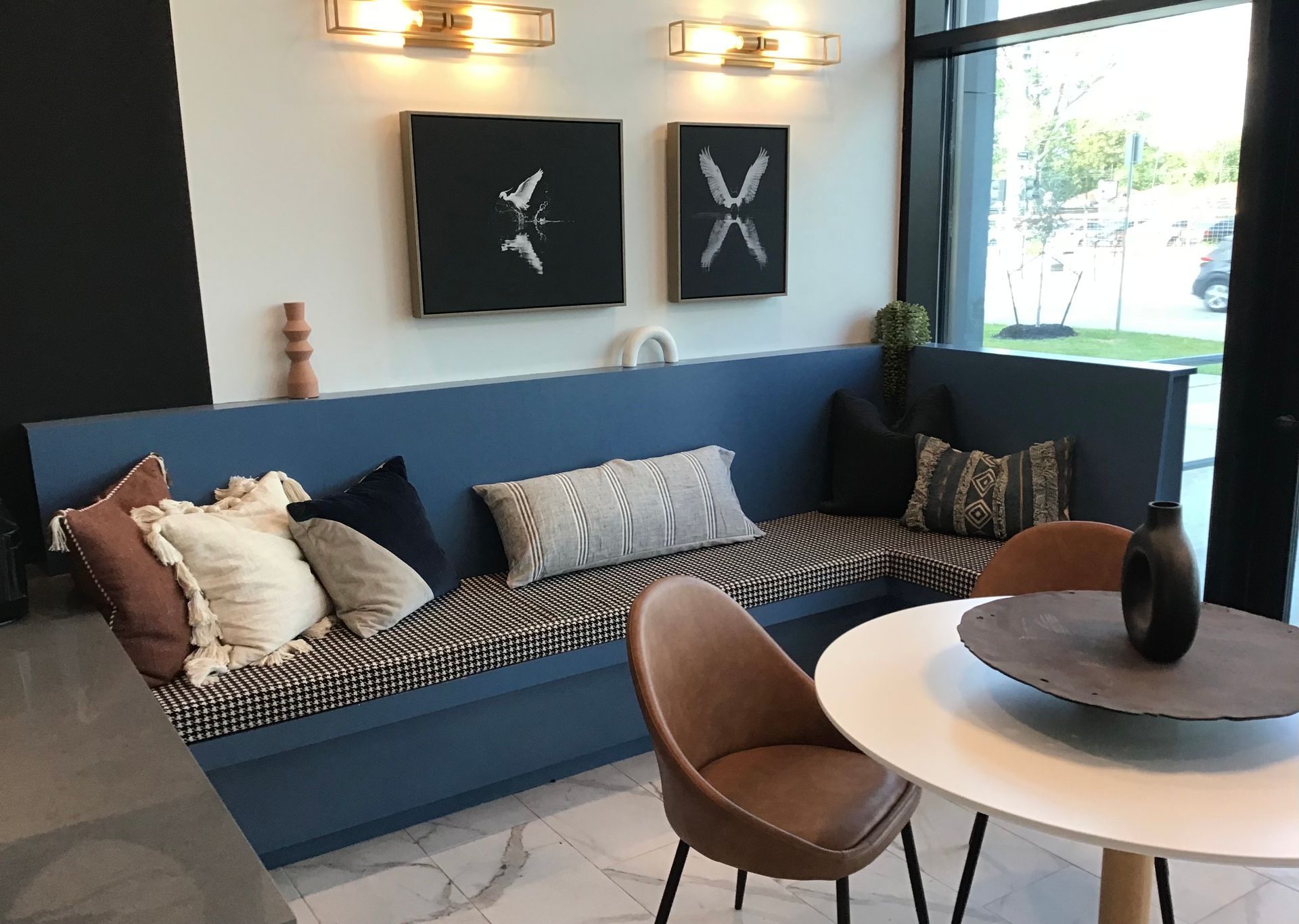 A blue corner booth with patterned cushions and pillows, paired with a small round table and brown chair in a room.