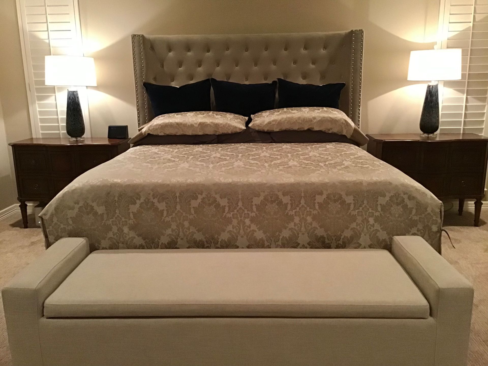 A bedroom with a tufted headboard, patterned comforter, two bedside tables with lamps, and a long upholstered bench.