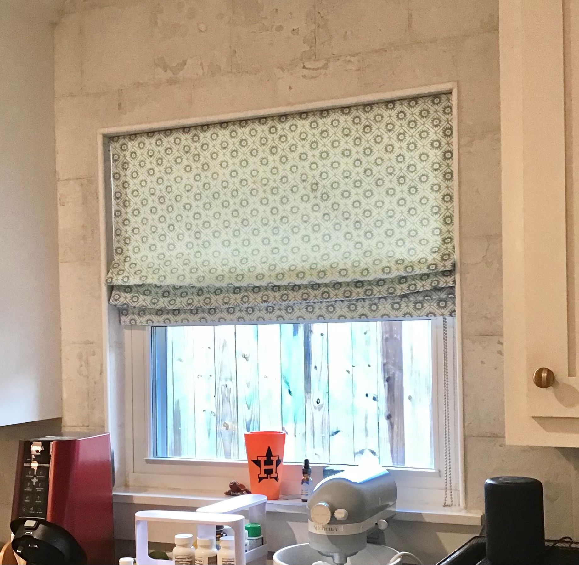A window with a patterned, green and white fabric Roman shade, set in a stone-textured wall in a kitchen.