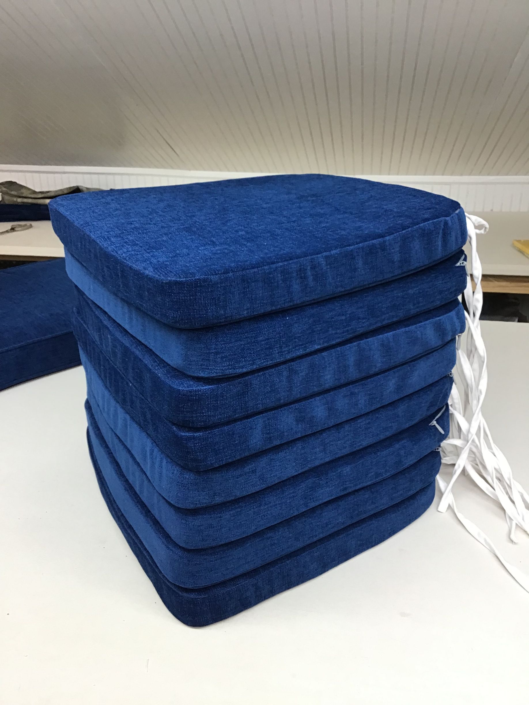 A stack of eight identical royal blue square chair cushions with white tie strings on the right, resting on a table.