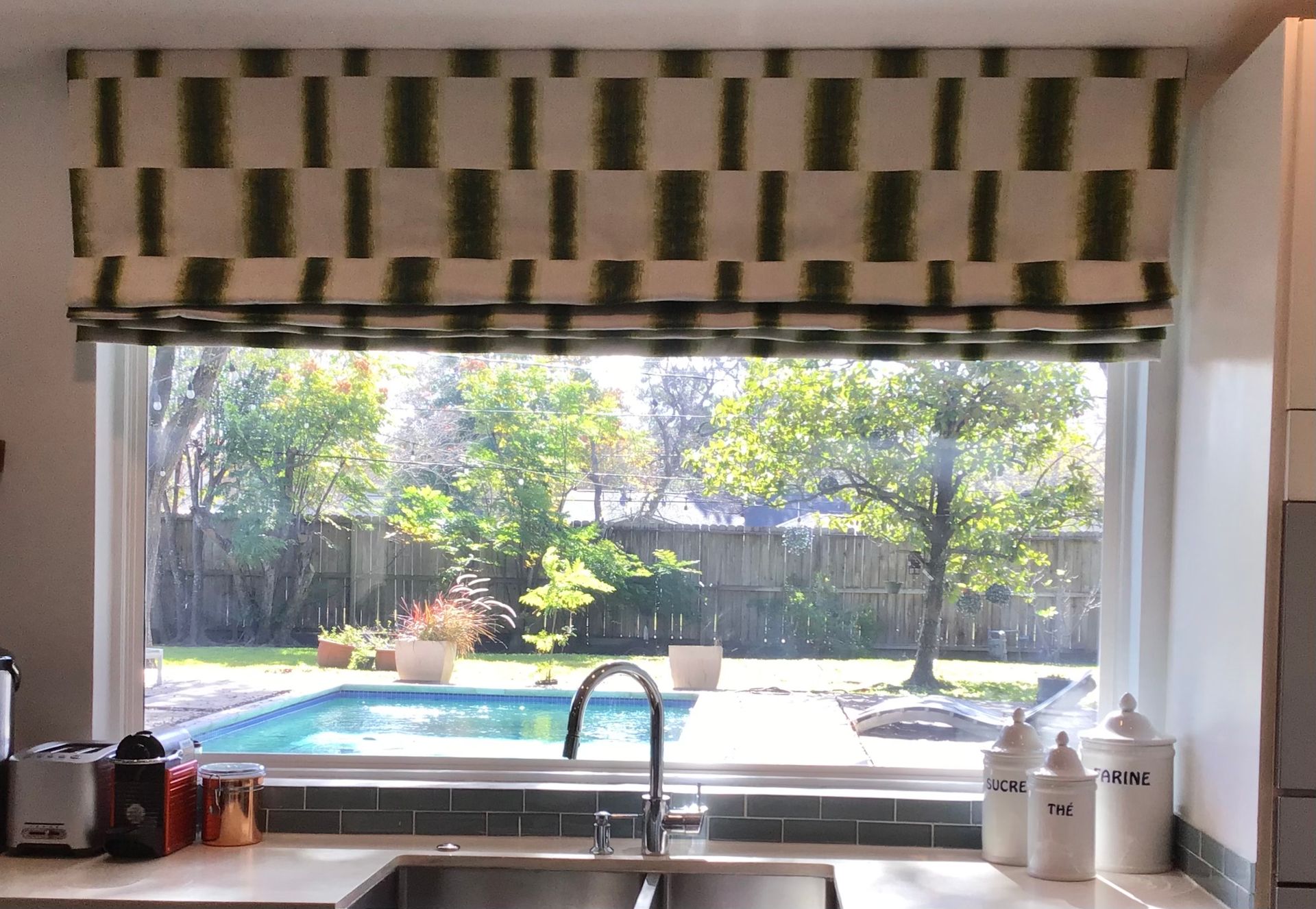 A kitchen window view looking out at a swimming pool, featuring a fabric Roman shade with a green and white pattern.