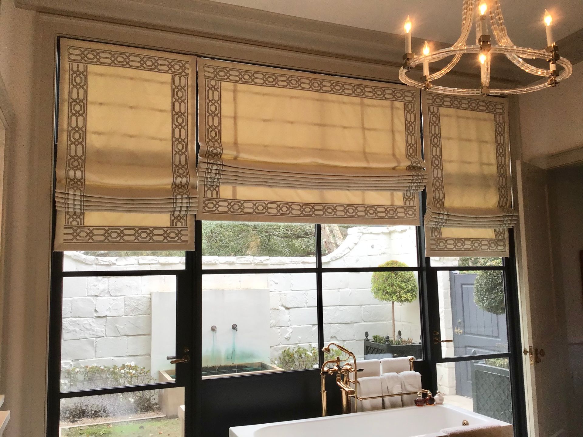 Three beige Roman shades with a geometric brown border hanging over large black-framed windows in a bright room.