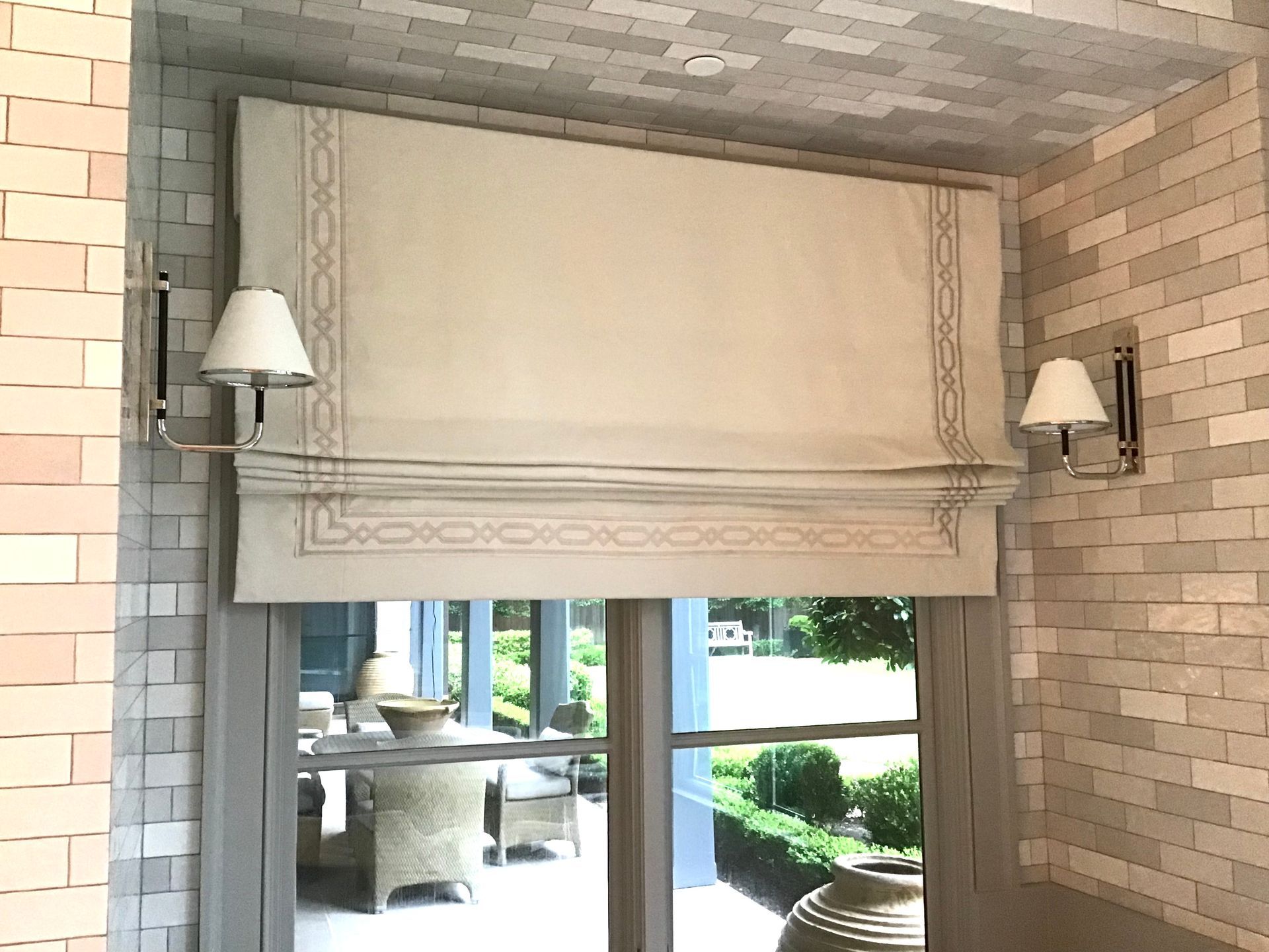 A fabric Roman shade with a decorative border hanging in a window between two wall-mounted lamps against brick walls.