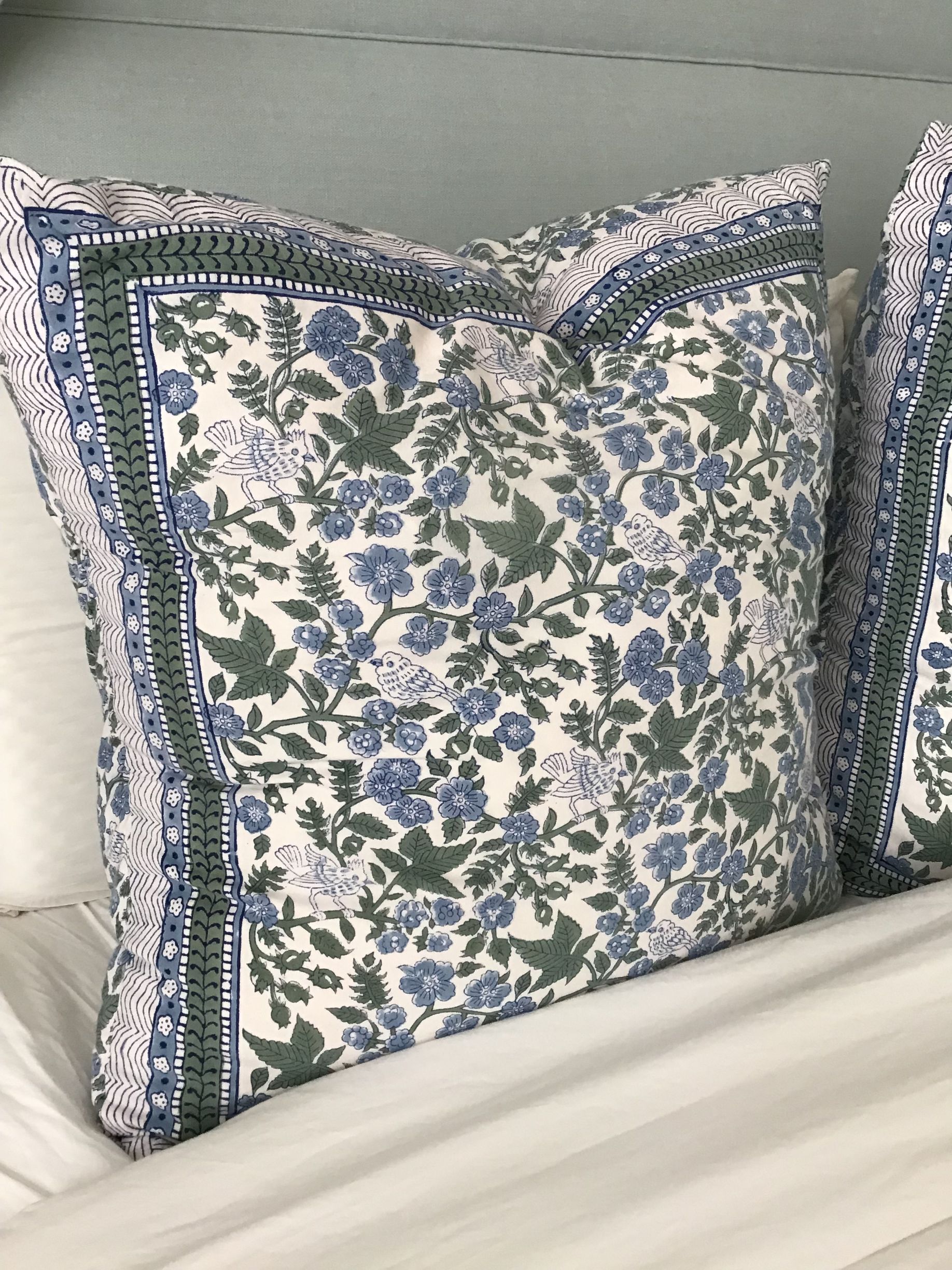 A decorative square pillow with a blue and sage green floral pattern and a dark green border, resting on a white bed.