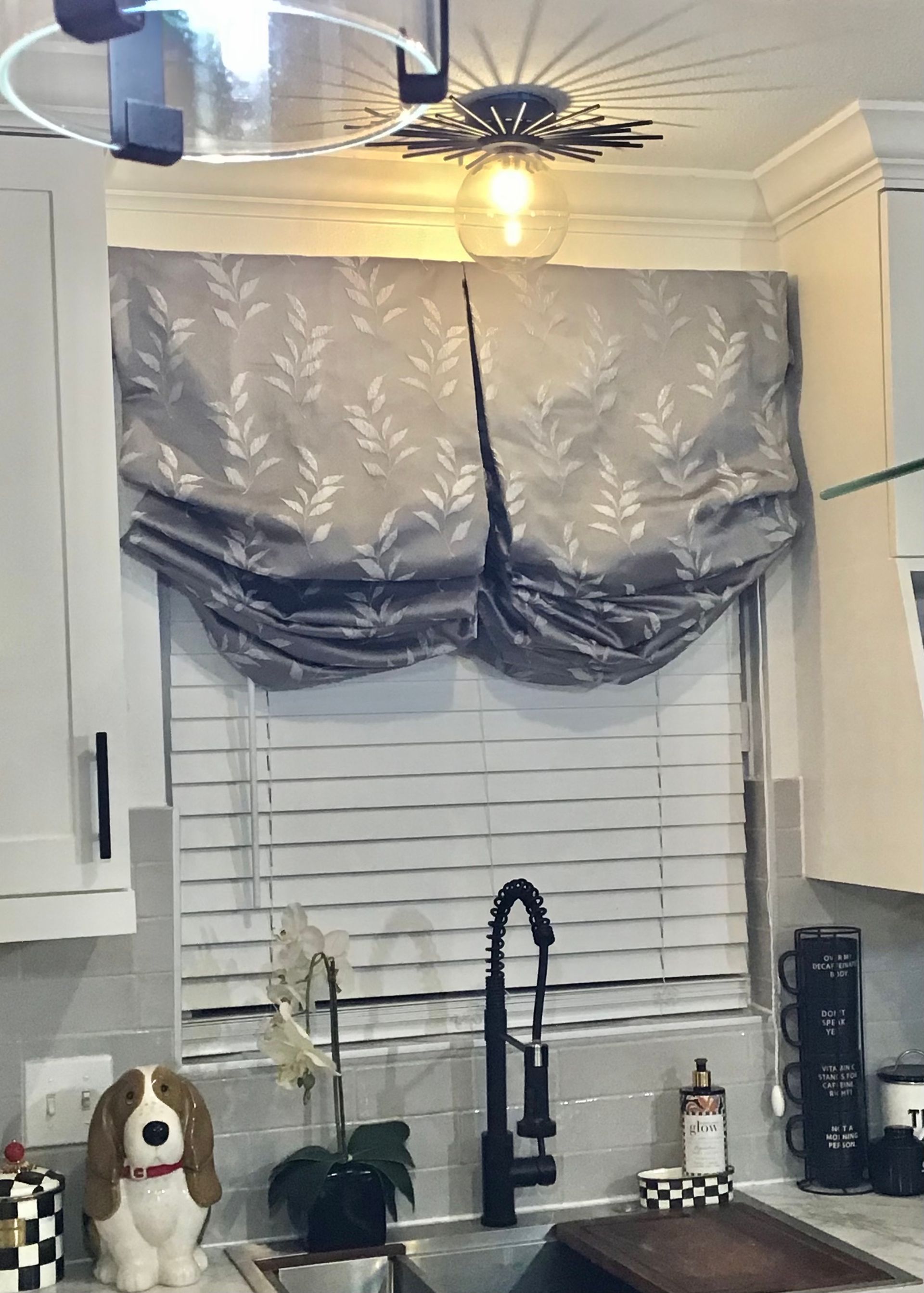 A kitchen window with grey leaf-patterned Roman shades above white blinds, featuring a modern light fixture and sink.