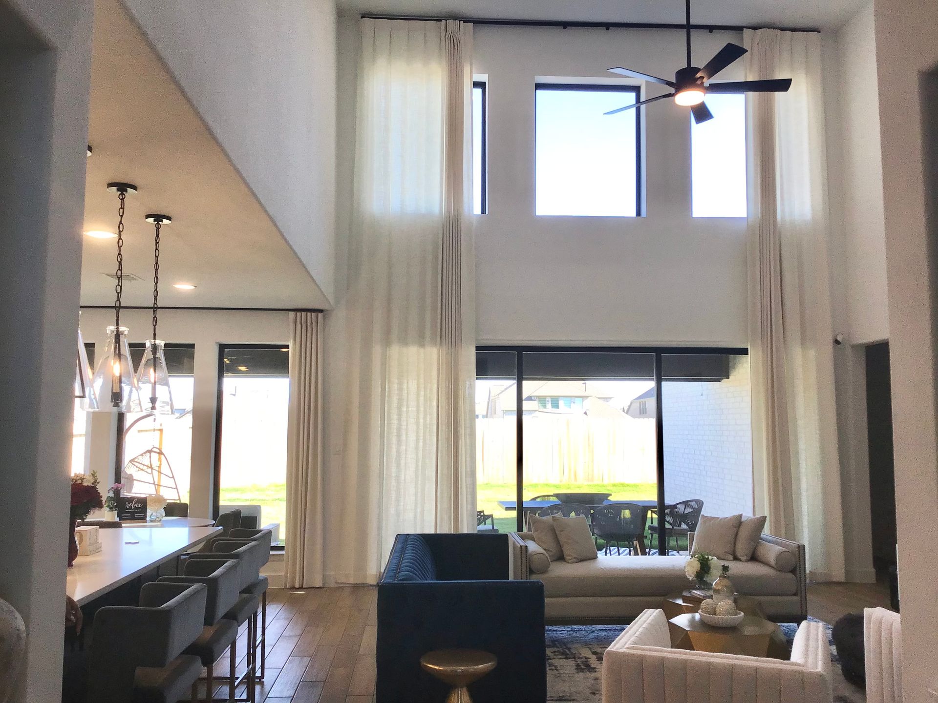 A bright, modern living room with tall windows, cream curtains, a dark blue sofa, and an open kitchen with bar seating.