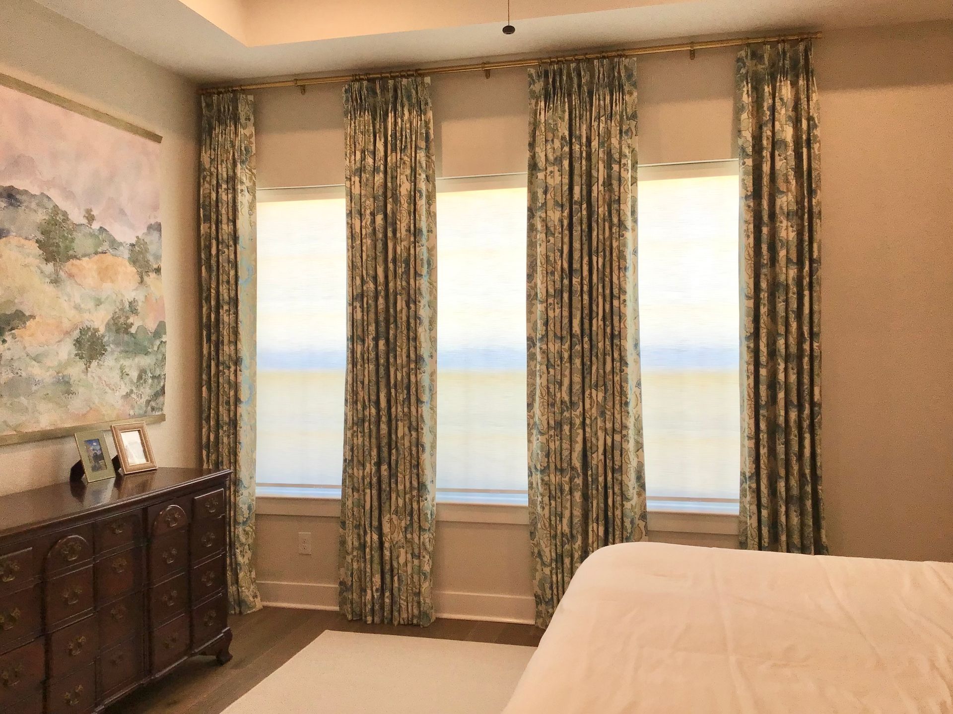 Floral curtains hang over large windows beside a dark wood dresser and a bed in a sunlit bedroom.