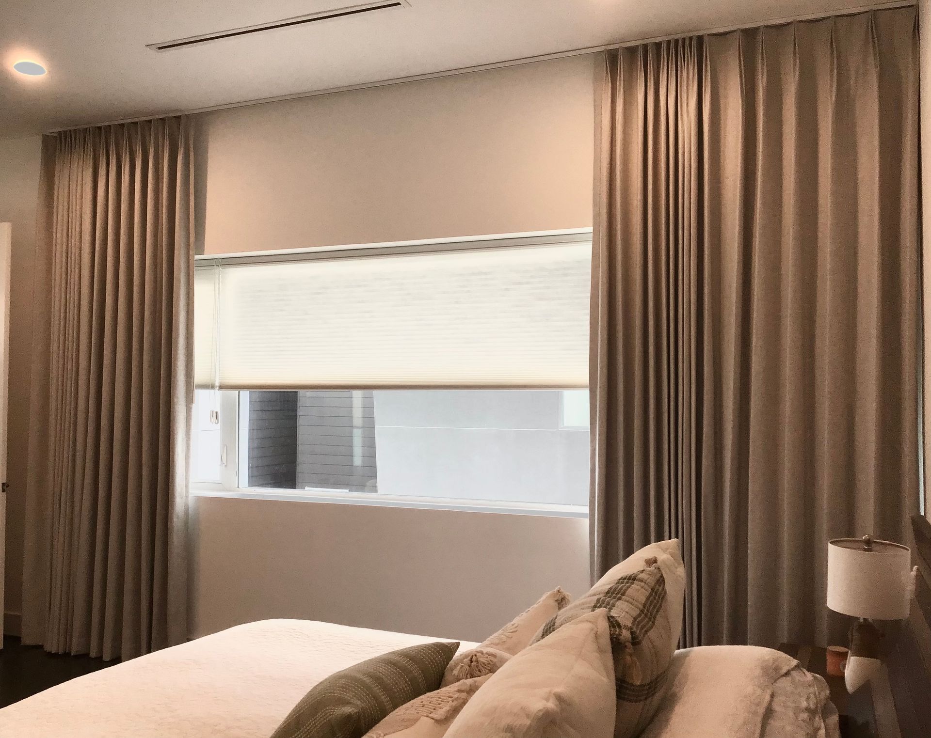 A bedroom featuring beige curtains flanking a window with a white shade, positioned above a bed with decorative pillows.