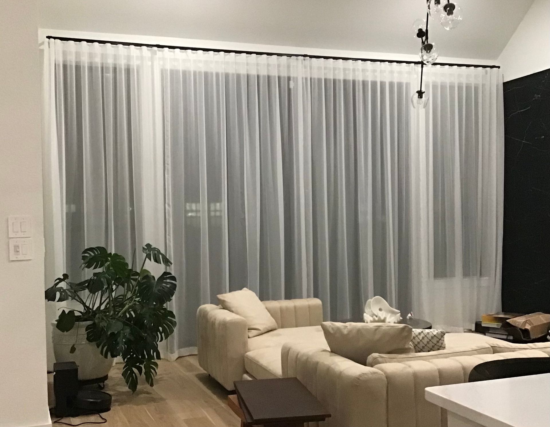 A living room with light-colored furniture, a large potted plant, and floor-to-ceiling sheer white curtains on a dark rod.