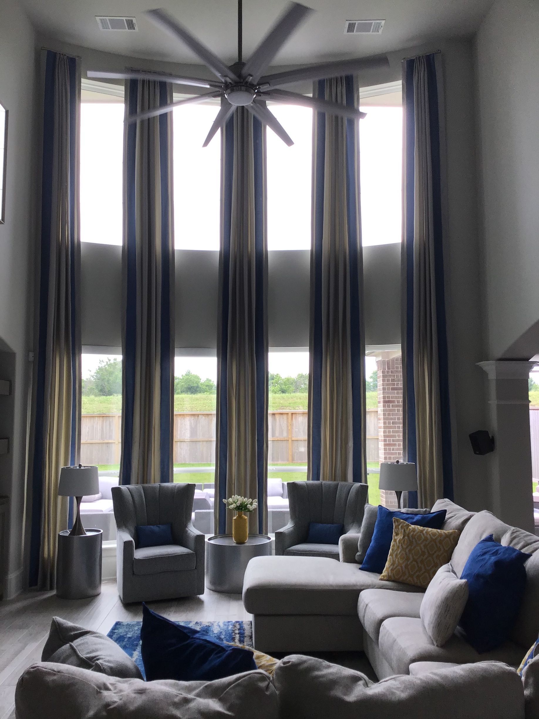 A living room with tall windows, vertical striped curtains, two gray armchairs, and a large beige sectional sofa.
