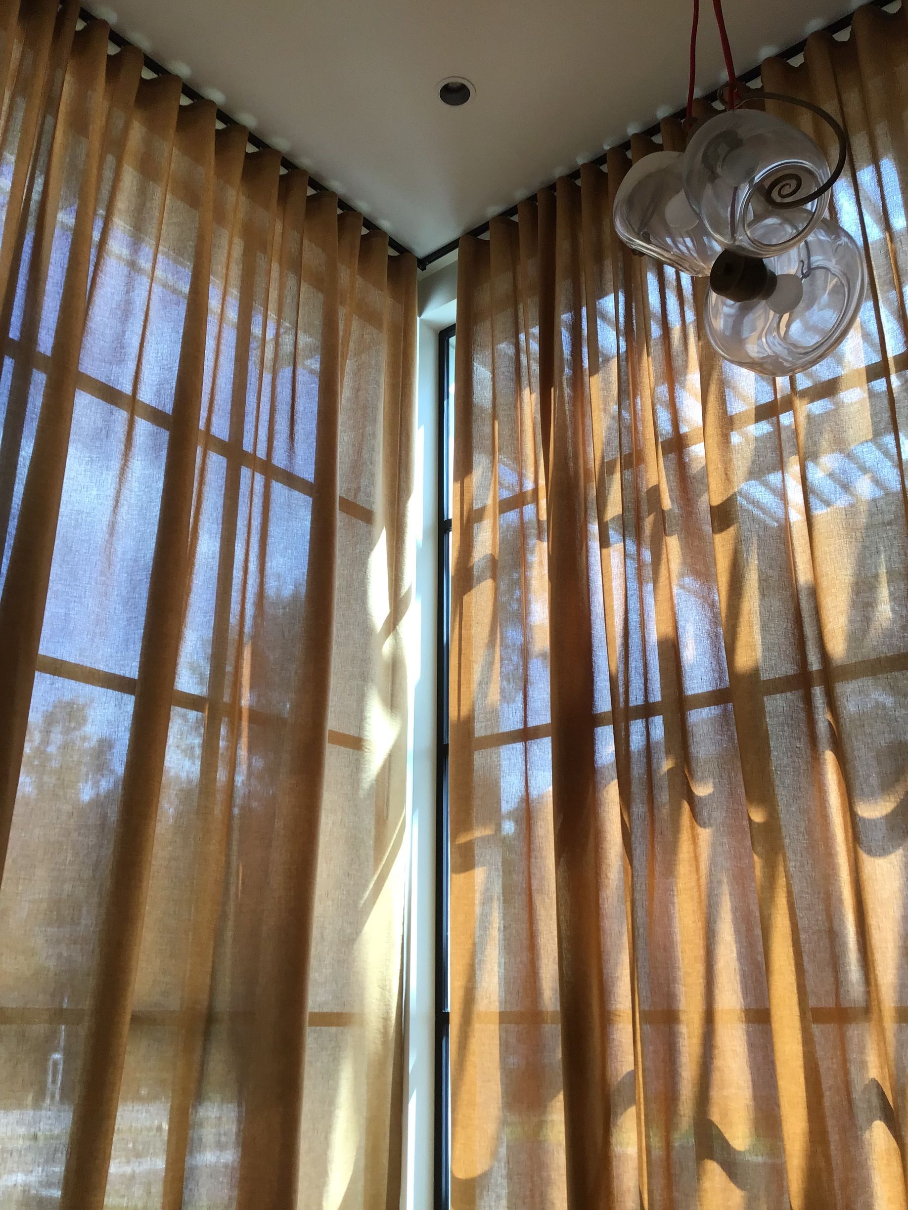 Floor-to-ceiling sheer brown curtains hanging in a corner, with light filtering through and a glass pendant light above.