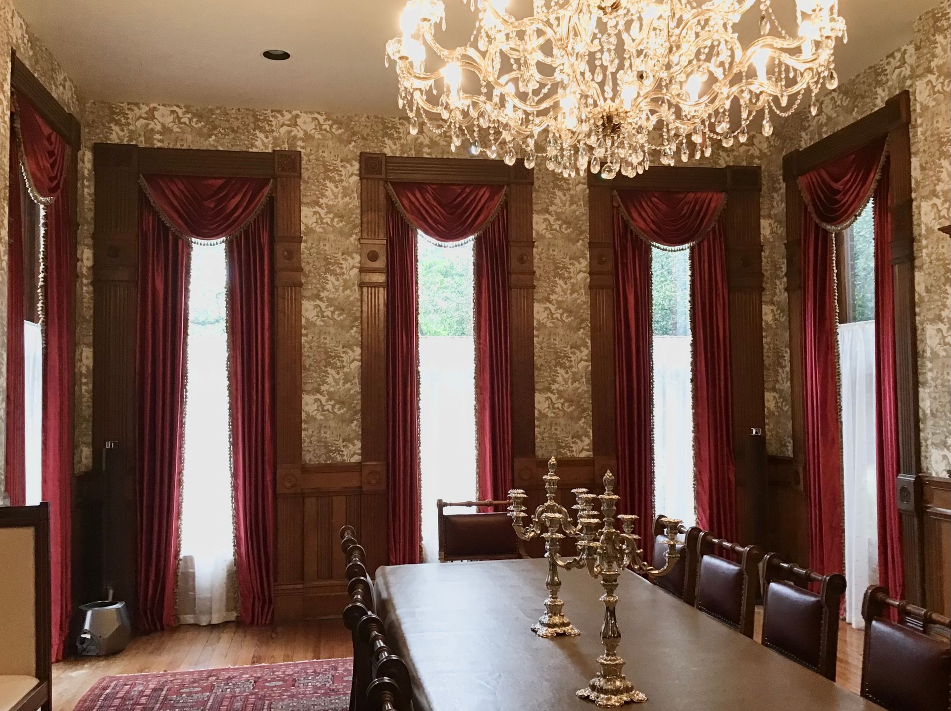 A dining room with a long table, gold candelabras, ornate red curtains, a crystal chandelier, and patterned wallpaper.