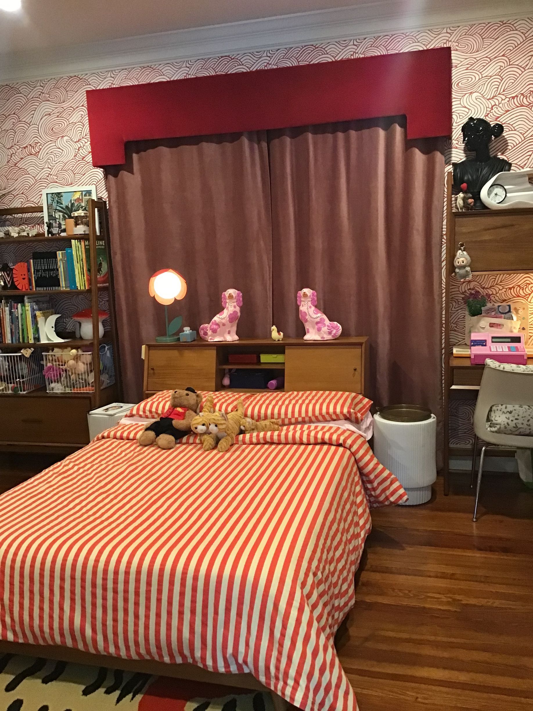 A bed with orange and white striped bedding, two pink figurines on a headboard, a window with red drapes, and a bookshelf.