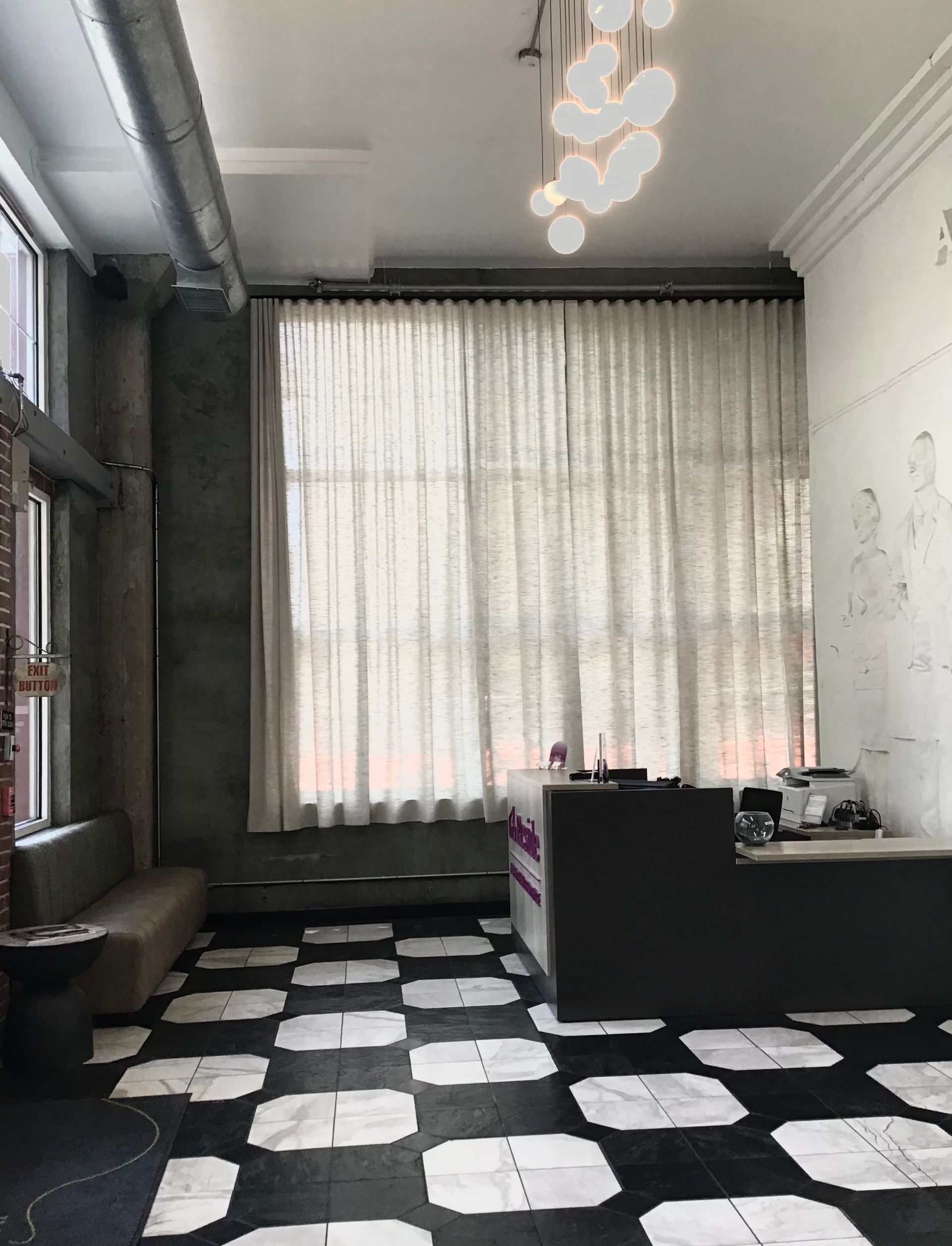 A reception area with a black and white tiled floor, a dark front desk, tall sheer curtains, and a modern light fixture.
