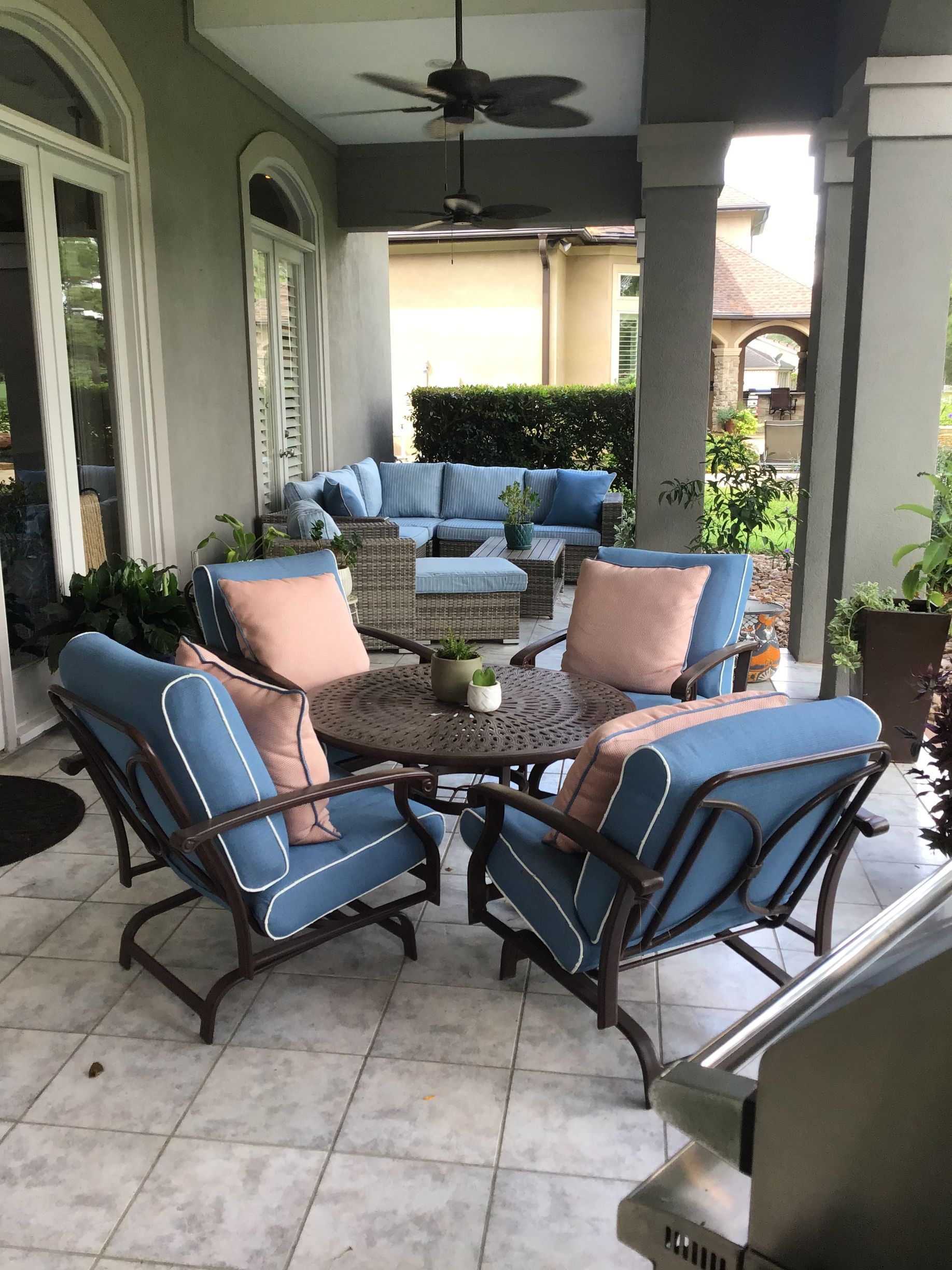 A patio set with four blue chairs and a round table on a stone floor, with a sectional sofa and greenery in the background.