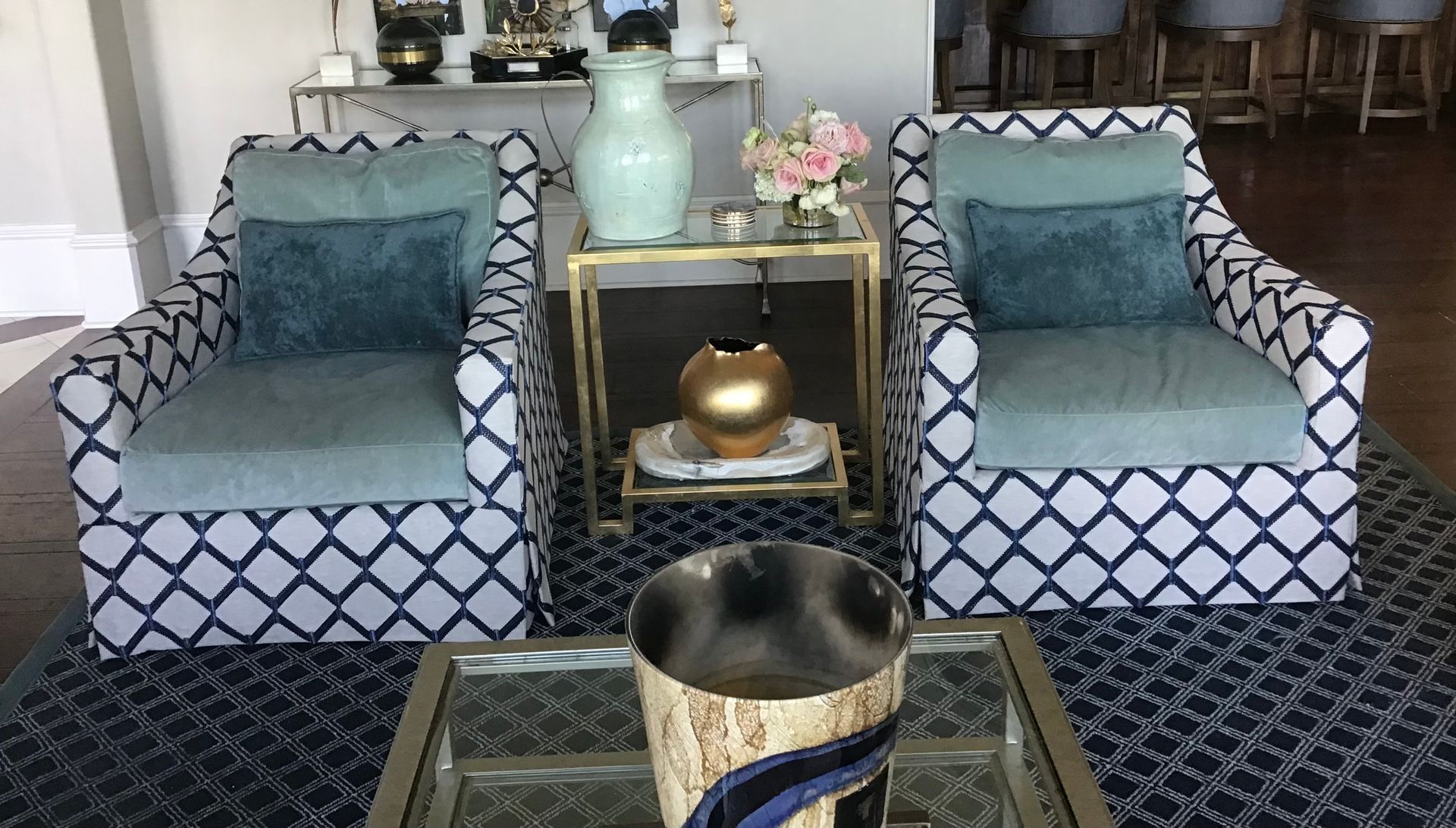 Two patterned armchairs with teal cushions sit on a blue rug, flanking a gold-trimmed end table with a vase and gold bowl.