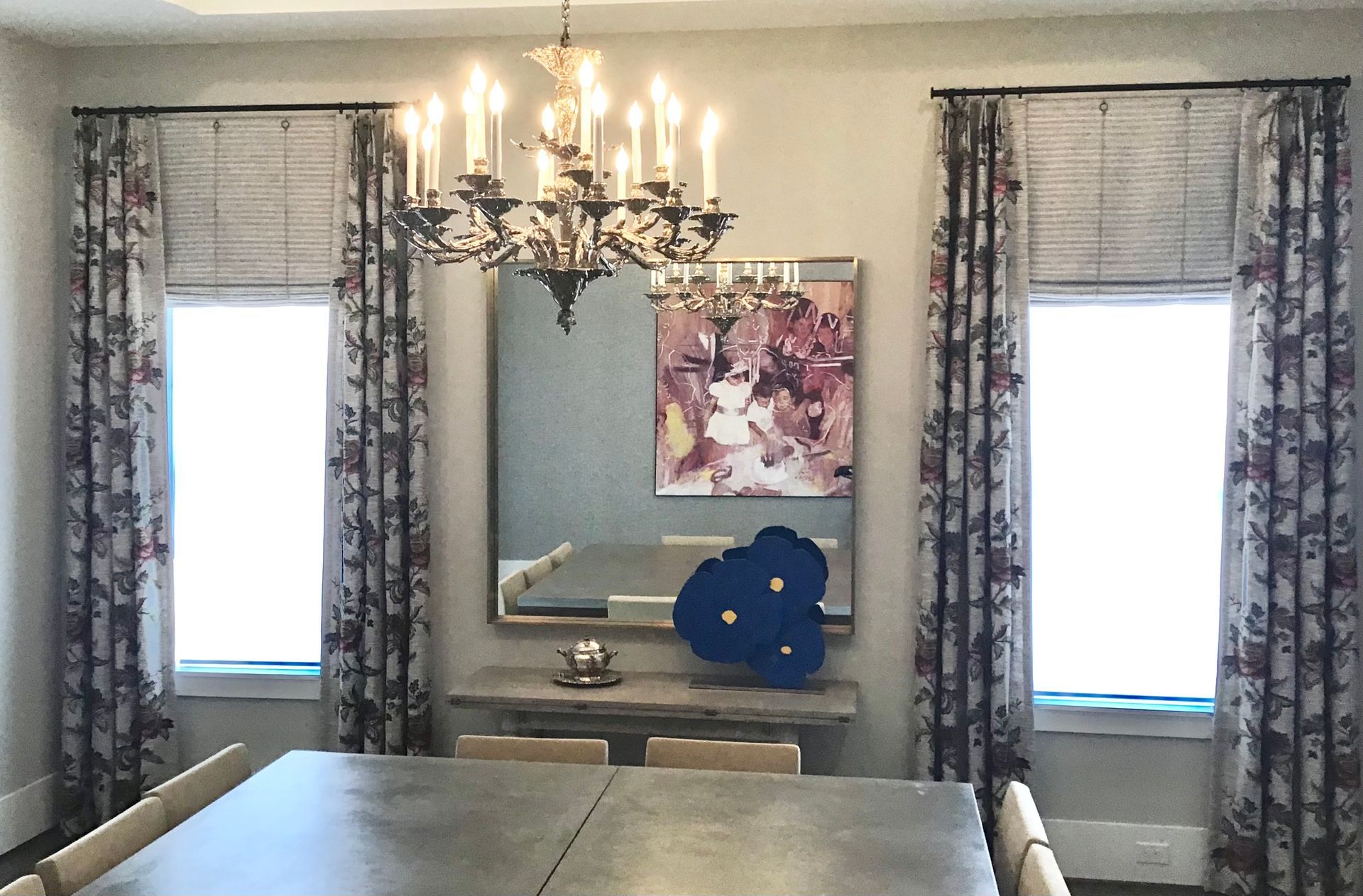 Dining room with a chandelier over a table, a central mirror with abstract art, and two windows with patterned drapes.