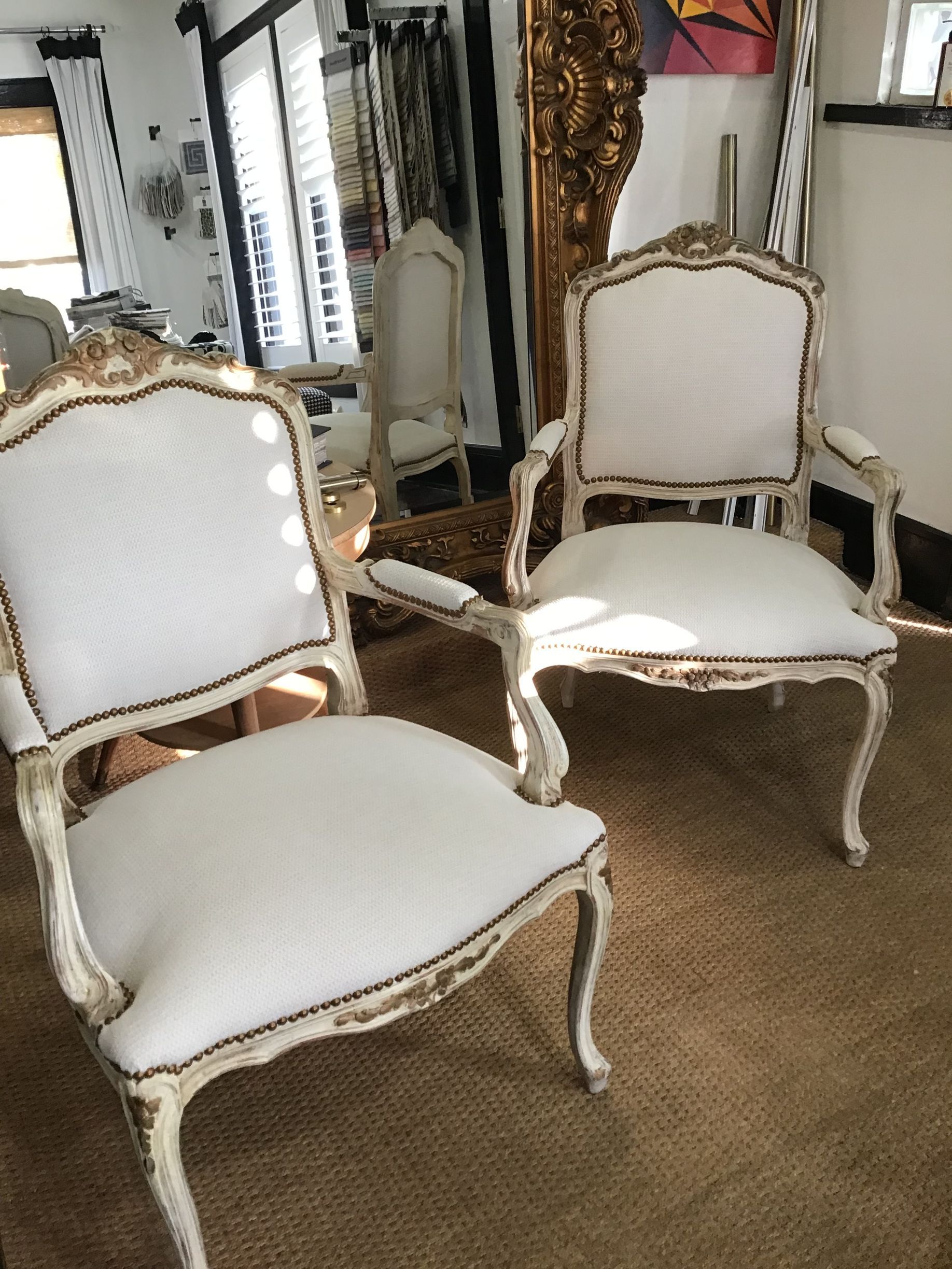 Two matching off-white armchairs with distressed wood frames sit on a brown rug in front of a large ornate gold mirror.