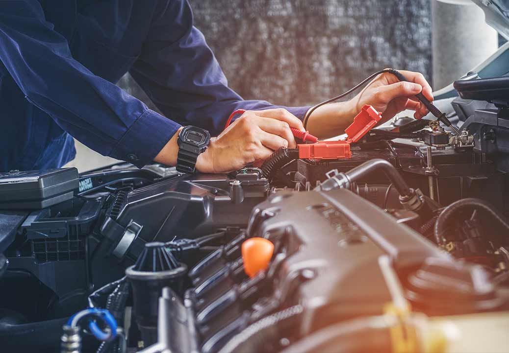 Mechanic in blue jumpsuit connecting jumper cables to a car battery in an engine bay.