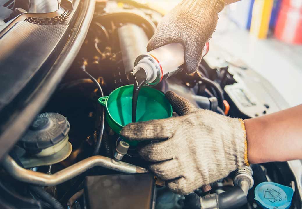 Mechanic pouring oil into a car engine using a green funnel, gloved hands visible.