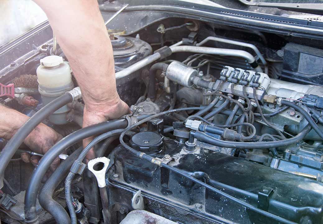 Hands working on a car engine, various hoses and components visible under the hood.